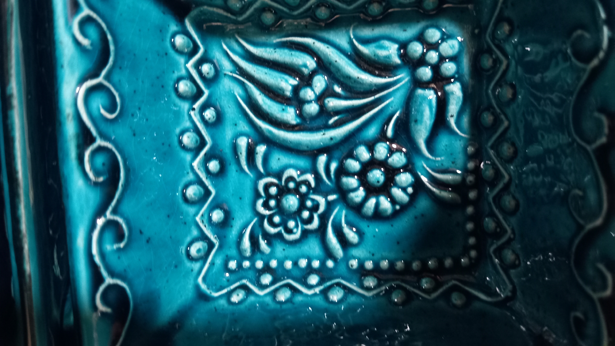 This is a close-up, detailed shot of an ornate, glazed ceramic tile or surface with a raised decorative pattern in shades of turquoise and teal. The design features stylized floral motifs, including what appear to be tulips and smaller, star-shaped flowers, interspersed with vine-like elements and small, raised dots. The surface is slightly glossy, with visible texture and some darker speckling within the glaze. The tile appears to be part of a larger object or installation. The image does not provide any contextual information about the city of Kos, Greece, or any environmental elements beyond the ceramic itself.