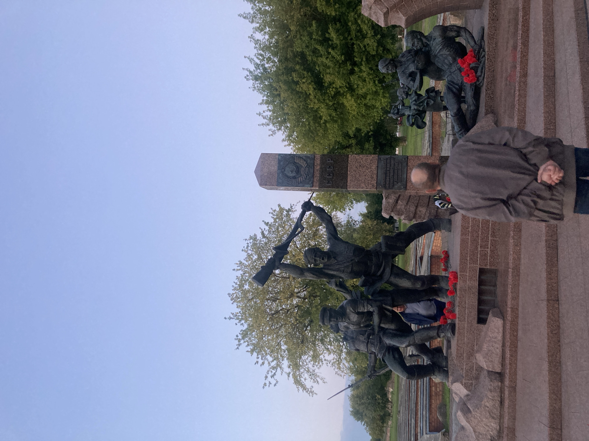 The image depicts the Hero-Defender of the Brest Fortress memorial complex, a significant historical site in Brest, Belarus. In the foreground, a man with a bald head and a gray jacket stands with his hands clasped behind his back, facing away from the camera, looking towards a large bronze monument.
The monument itself is a multi-figure sculpture depicting soldiers in combat. One prominent figure holds a rifle above his head, while others are positioned in dynamic poses, suggesting struggle and resistance. Behind the soldier figures, a tall granite obelisk stands, bearing Soviet symbols and inscriptions. Red carnations, a traditional symbol of remembrance in Belarus, are placed at the base of the monument.
To the right of the main sculpture, another bronze depiction shows figures, including a fallen soldier and women, further emphasizing the theme of sacrifice and resilience. Red carnations are also present here. The background shows lush green trees and a clear sky, indicating an outdoor setting. The overall scene conveys a somber and respectful atmosphere, characteristic of a war memorial.