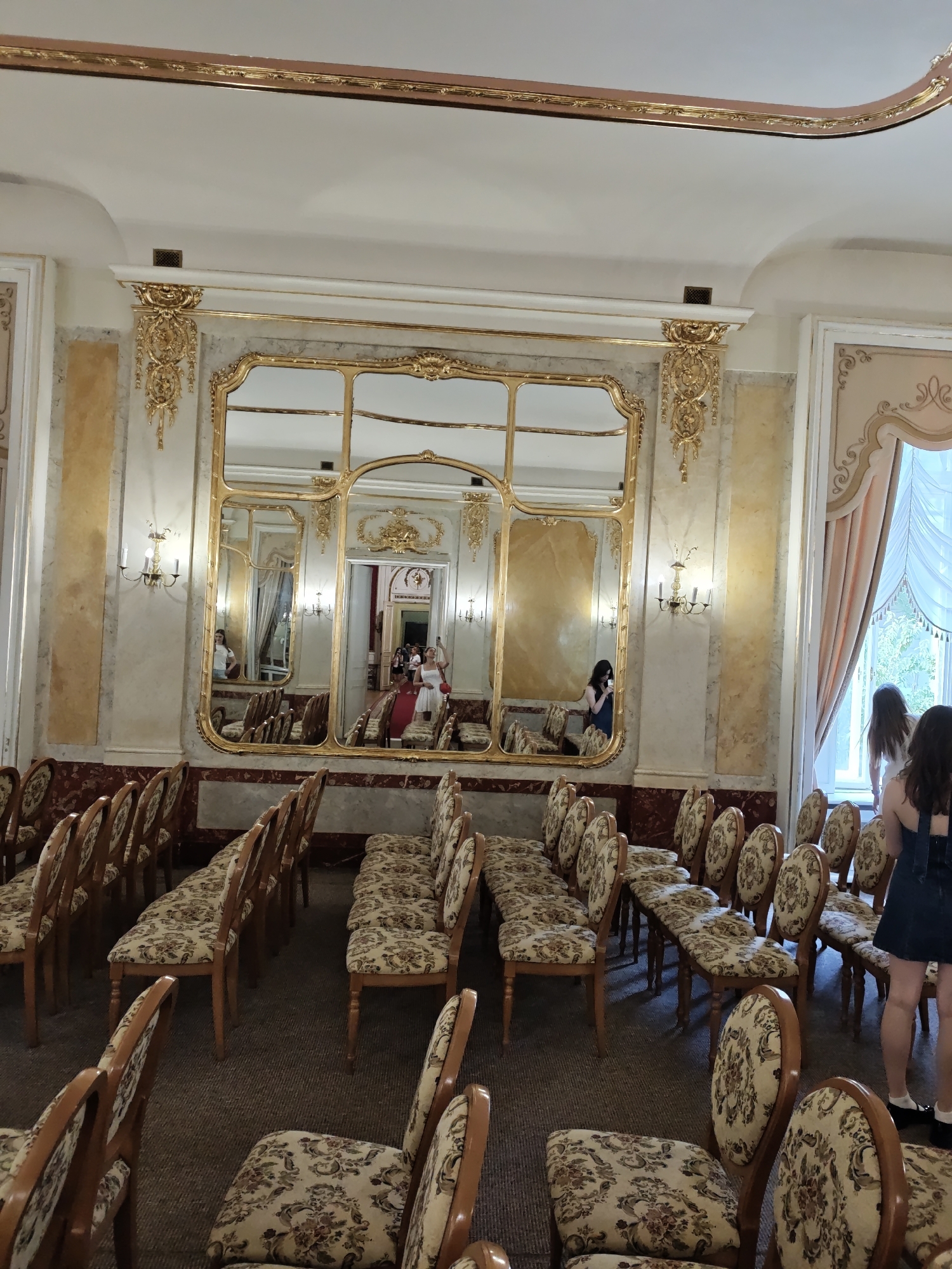 A wide-angle interior shot captures a richly decorated room likely serving as a venue for events or performances. The room is furnished with rows of upholstered chairs, their seats and backs adorned with a floral pattern in muted tones of beige, brown, and red. The chairs have wooden frames, some with ornate carvings.
Dominating the back wall is a large, ornate, gilded mirror with a multi-panel design, reflecting the interior of the room. The mirror is framed by elaborate gold scrolling and decorative elements. On either side of the mirror, wall sconces with multiple candle-like lights are mounted. The walls themselves are a mix of marble-like paneling in beige and gold, with sections of darker patterned wallpaper or fabric near the floor.
To the right, a large window is dressed with draped, light-colored curtains, and beyond it, greenery suggests an outdoor view. Several individuals are visible in the scene. In the reflections of the mirror, people are seen standing and moving. Two women are also visible in the foreground on the right, one wearing a dark denim dress and the other in a white top, with their backs to the camera, appearing to be near the window. A woman in a white dress is also visible in the reflection of the mirror, seemingly posing.
The ceiling is high and features decorative molding, also highlighted with gold accents. The flooring is carpeted in a dark, neutral color. The overall impression is one of opulence and historical grandeur.
This room is located in the Lviv National Opera House in Lviv, Ukraine. This specific area appears to be an anteroom or a waiting area adjacent to a performance hall, designed to impress visitors with its elaborate decor.