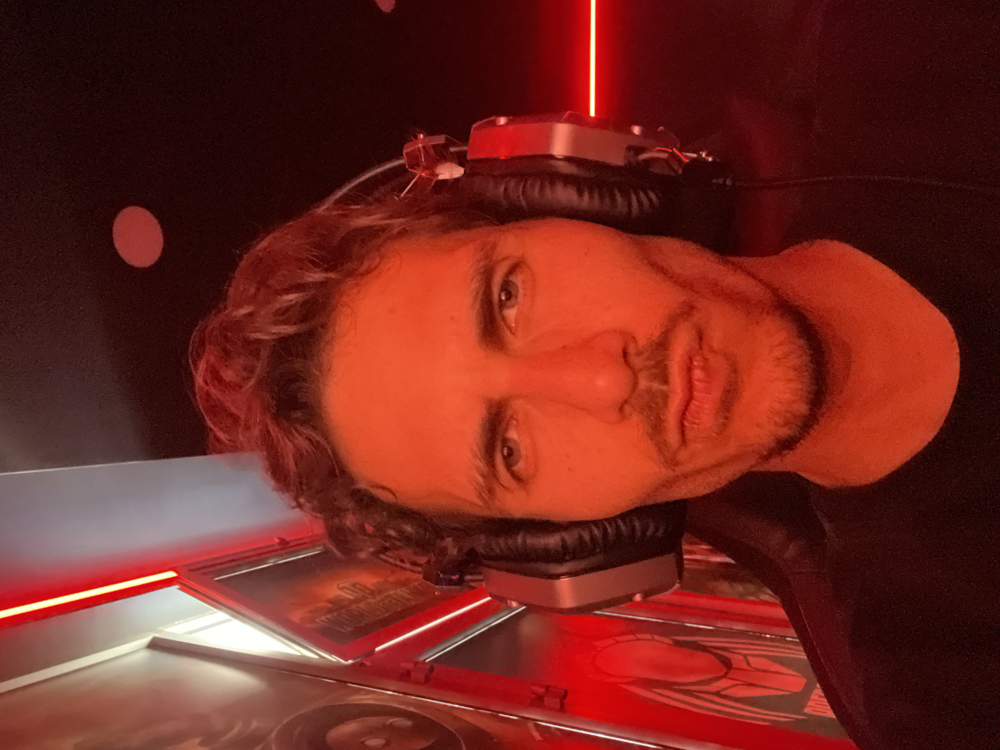 A man with curly brown hair and a light beard is wearing headphones. He is in a dark room lit with red light. In the background, there is a red light strip and a red circle. There is a table or display in front of him with images on it, also lit with red light. The image is taken at an angle, with the man's head tilted to the side. There is no information in the image to determine that this image was taken in Budva, Montenegro.