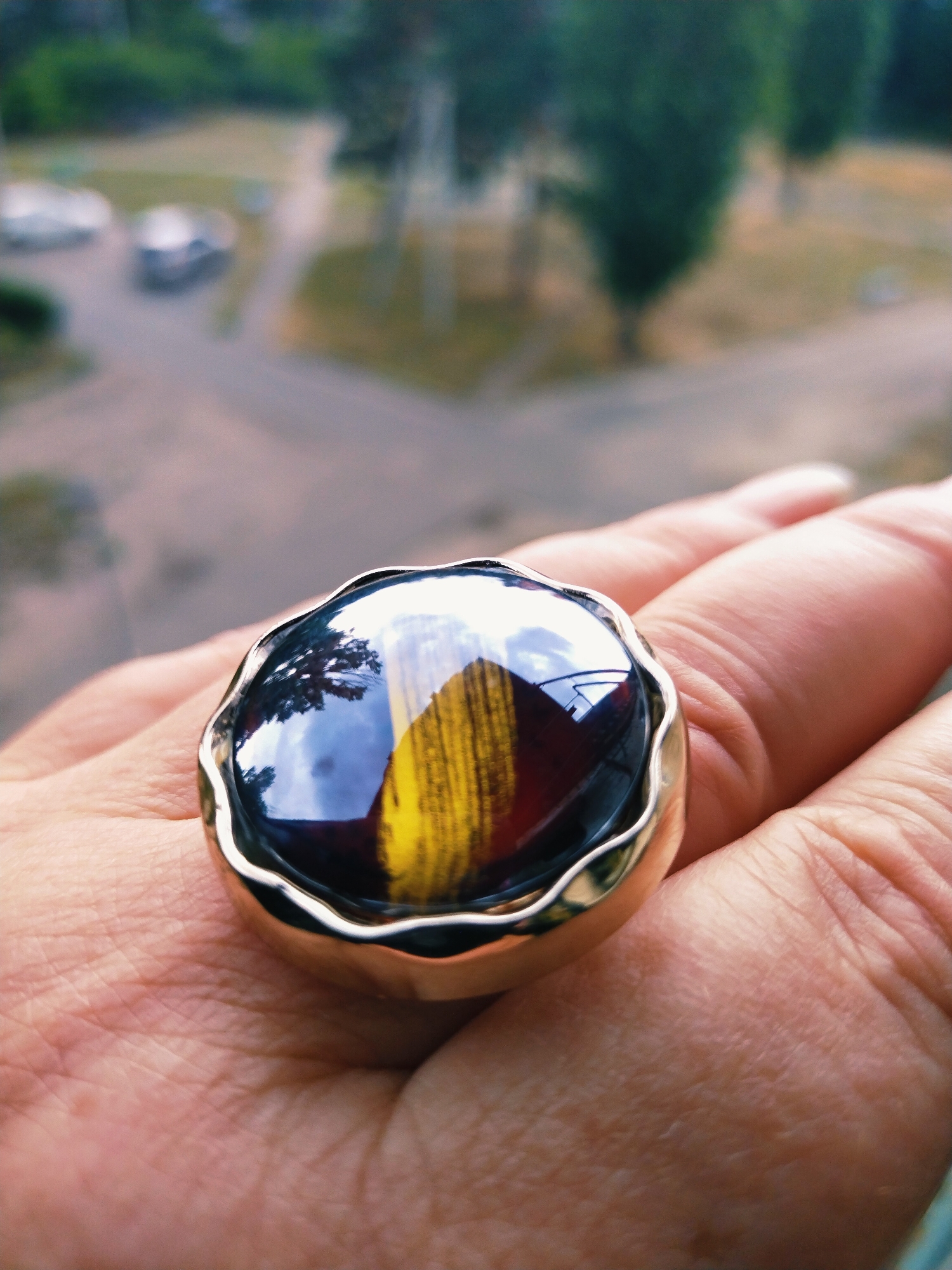 The image displays a close-up of a ring on a person's hand, with a blurred background. The ring features a large, multi-colored stone with reflections. The hand appears to be resting on a surface, and the skin texture is visible. The background shows a street scene with cars and greenery, suggesting an outdoor urban environment. The exact location within Horishni Plavni, Ukraine, cannot be determined from the provided image.