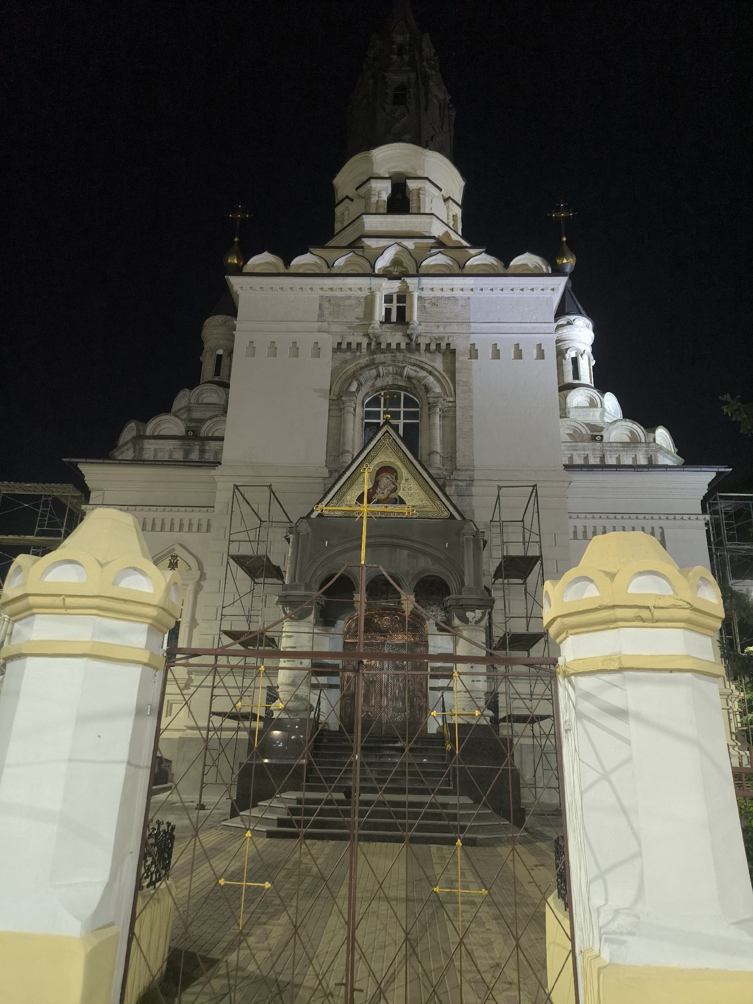The image shows the exterior of the Saint Nicholas Cathedral in Mykolaiv, Ukraine, at night. The cathedral is illuminated and features a white facade with ornate architectural details, including domes and crosses. Scaffolding is visible on the front of the building, suggesting ongoing renovation or construction. In the foreground, a decorative fence with yellow crosses and a gate partially obscures the entrance. The sky is dark, indicating it is nighttime. There are no people visible in the image.