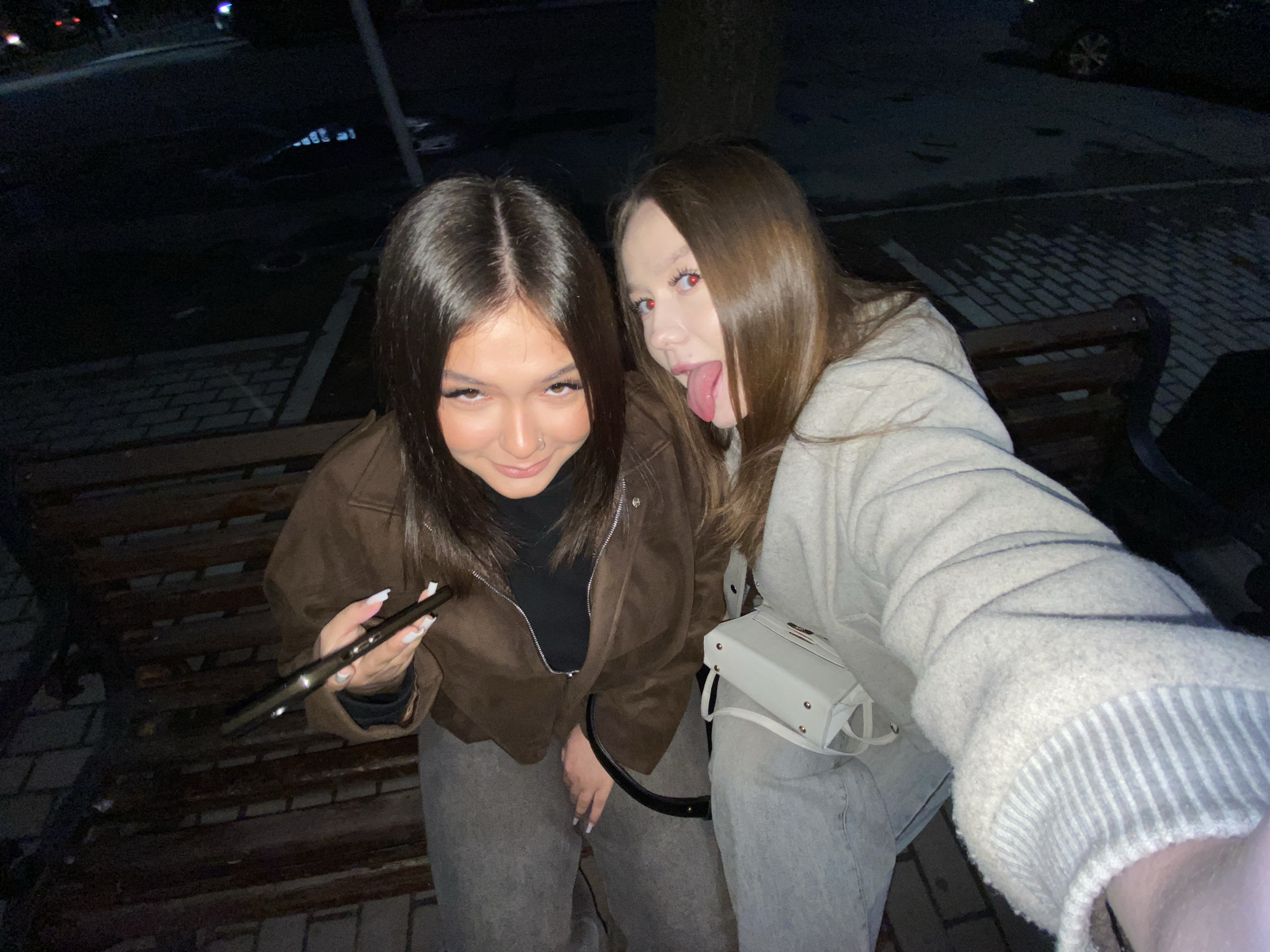 A close-up, flash-lit photo taken at night in Chaikovsky, Russia, features two young women posing playfully. The woman on the left has dark hair, a nose piercing, and is wearing a brown zippered jacket over a black top and gray pants. She looks towards the camera with a slight smirk and holds a slender, dark object, likely an electronic vaping device, in her left hand, revealing manicured nails with white tips. The woman on the right, with lighter brown hair, is wearing a light-colored, possibly off-white or light gray, fuzzy jacket and gray pants. She is seen sticking out her tongue in a playful expression, and both her eyes, like those of the other woman, show a prominent red-eye effect from the flash. A small white shoulder bag hangs from her side. They are seated on a dark wooden park bench. The background is mostly obscured by darkness, with faint outlines of paved ground, possibly cobblestone, and indistinct elements of an outdoor urban setting, including what appears to be a curb and the very faint outline of a parked vehicle. No visible text is present in the image.