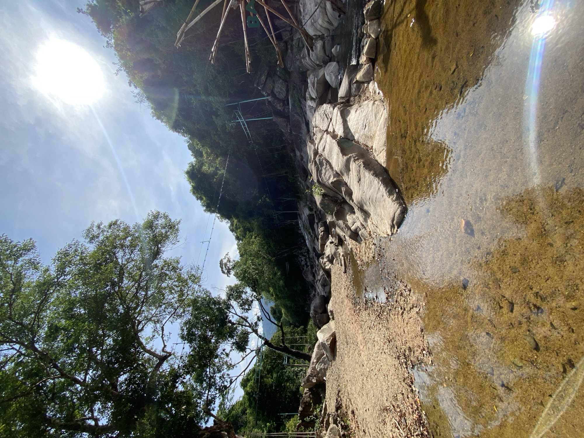 The image depicts a natural outdoor scene, likely a stream or shallow riverbed, characterized by rocky terrain and lush vegetation under a bright, sunny sky. The foreground features a sandy, dried-out area with scattered leaves, transitioning into a shallow, clear body of water with a rocky bottom. Large, weathered rocks are prominent throughout the scene, with some forming a natural pathway or structure in the water.
The midground reveals more substantial rock formations and a densely wooded hillside forming the backdrop. The vegetation consists of various trees and plants, suggesting a tropical or subtropical environment. The presence of what appear to be man-made structures, such as poles and cables stretching across the scene and a ladder-like structure on the hillside, indicates some level of human activity or infrastructure in the area, possibly related to a park, resort, or recreational facility. In the far right, part of a large, rustic wooden structure with bamboo elements is visible, resembling a part of a water wheel or decorative feature.
The sky is a bright blue with a few wispy clouds, and the sun is shining intensely, creating lens flare effects in the water and sky. The overall impression is a serene, somewhat untamed natural landscape with touches of human intervention. While the prompt mentions Hoa Thang Ward, Vietnam, this specific detail cannot be confirmed solely from the visual information presented in the image.