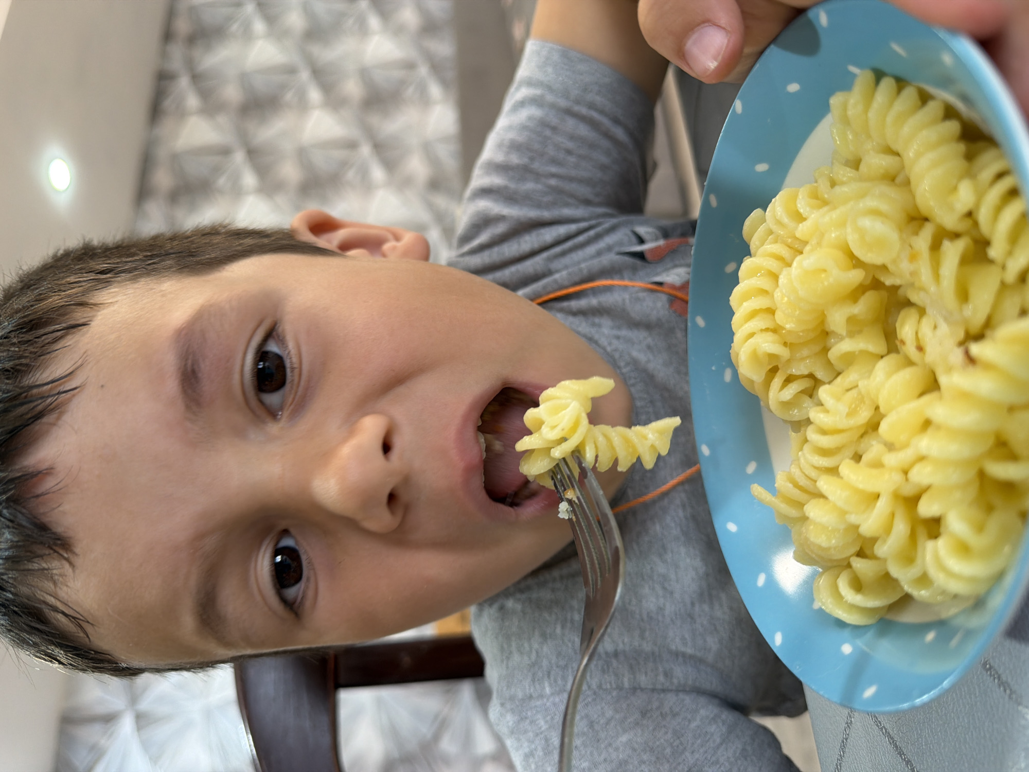 A close-up shot shows a young boy eating pasta from a blue and white polka-dotted bowl with a fork. The boy, who appears to be around 5-7 years old, is wearing a grey t-shirt and has dark hair. He is in the process of bringing a forkful of fusilli pasta to his open mouth. The pasta appears to be coated in a light sauce. The background is out of focus, but hints at a dining setting with a patterned wall and a chair. The lighting suggests an indoor environment, possibly a home. The boy's expression is one of focused enjoyment as he eats.