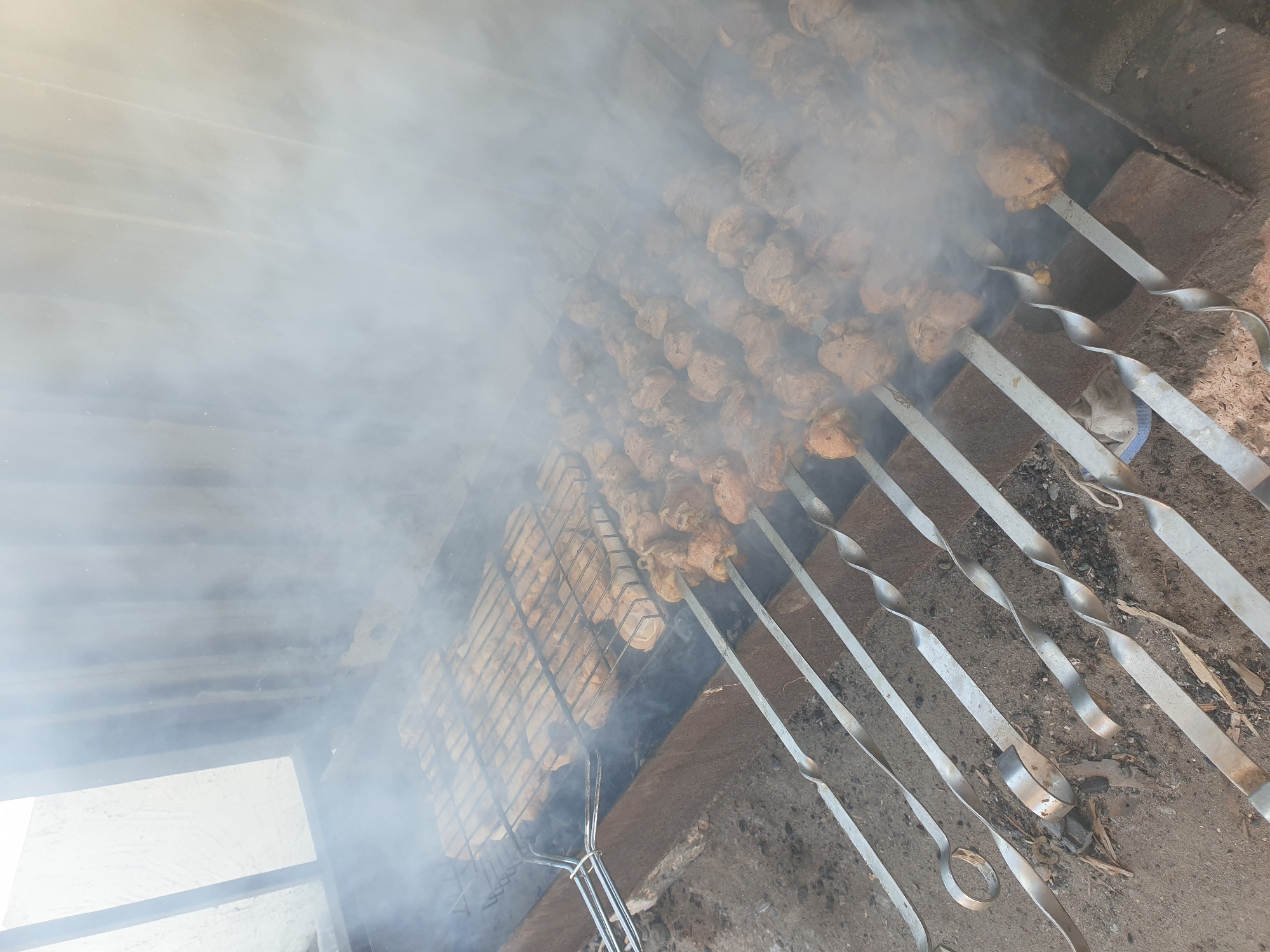 The image depicts an outdoor barbecue scene with numerous skewers of meat cooking over coals, producing a significant amount of smoke. In the foreground, several metal skewers with spiral handles are arranged horizontally, each holding pieces of seasoned meat. Behind these, a metal grill basket is filled with more meat, appearing to be chicken or pork, also undergoing the cooking process. The barbecue pit itself is constructed from brick or concrete. The background shows a corrugated metal structure, suggesting an outdoor shelter or enclosure. The ground beneath the barbecue is dirt and ash, indicative of a fire pit area. The scene appears to be located in Kuryk, Kazakhstan, a coastal city known for its fishing and port activities. The image captures a moment of food preparation, likely a gathering or meal being enjoyed outdoors.