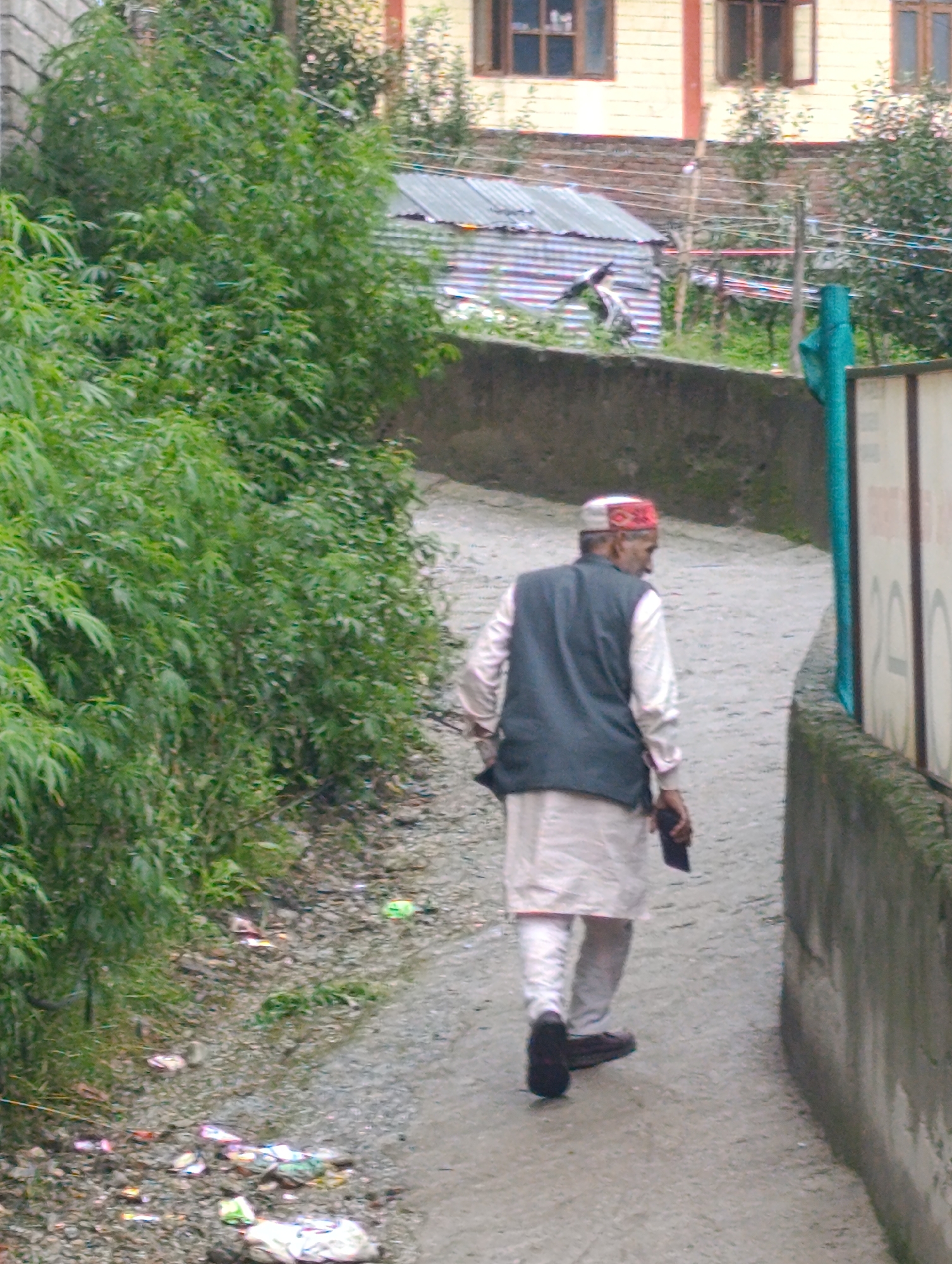 In the city of Aleo, India, a man walks away from the camera on a narrow, unpaved path. He is wearing a white tunic and trousers, a dark vest, and a red and white patterned cap. He is carrying a dark object in his right hand. The path is bordered on the left by dense green foliage and on the right by a concrete wall. The ground is littered with colorful debris. In the background, buildings with corrugated metal roofs and windows are visible.