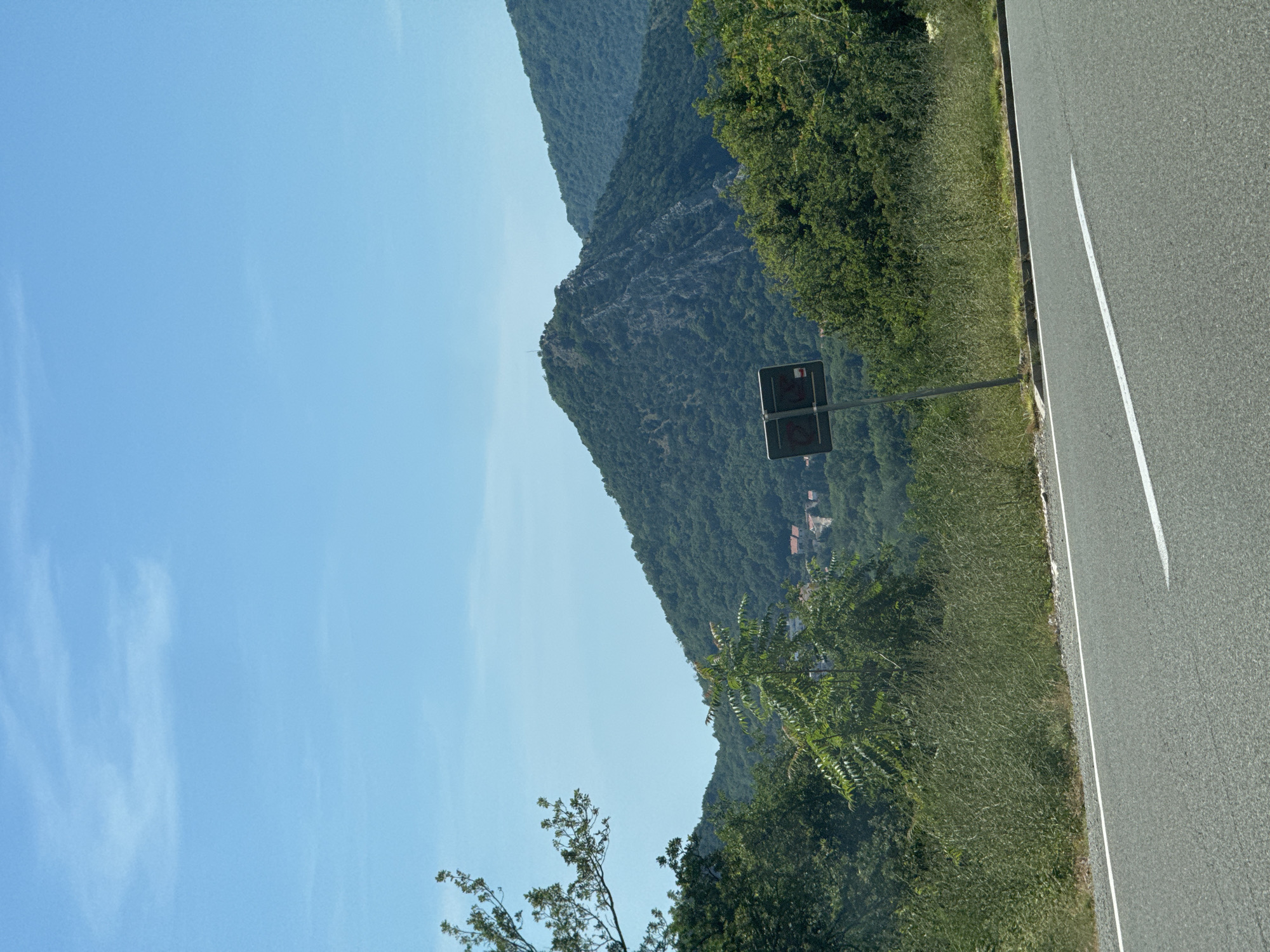 The image depicts a scenic view of a mountainous landscape near Golubovići, Montenegro. A paved road with a white lane marker runs diagonally across the lower part of the frame, suggesting a roadside perspective. To the left and right of the road, lush green vegetation and trees create a dense natural border.
In the middle ground, a prominent, tree-covered mountain dominates the scene. Its rocky peak is visible, and a small structure, possibly a tower or monument, can be seen at its summit. The slopes of this mountain are densely forested with various shades of green foliage. Further in the background, rolling hills covered in similar vegetation recede into the distance, culminating in a range of larger mountains. A small cluster of buildings, identifiable by their red rooftops, is nestled within the valley on the slope of one of the mountains, hinting at a settlement or village.
A road sign, featuring a dark rectangular panel with a red symbol and a white arrow, is positioned on a metal pole in the foreground, to the left of the central mountain. The sky above is a clear, bright blue with a few wispy clouds scattered across it, indicating a sunny day. The overall impression is one of natural beauty and tranquility, with the road and sign suggesting human presence within a natural environment.