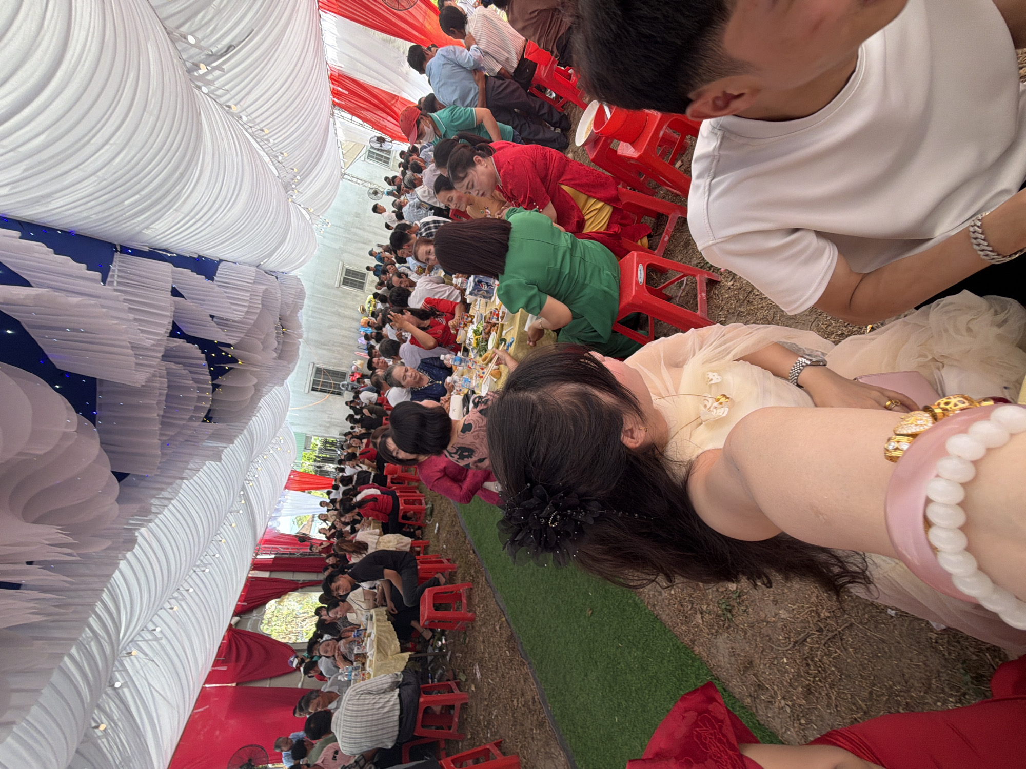 A high-angle view of a seated gathering of people under a tent-like structure in Tay Son Commune, Vietnam. The tent has white fabric draping and red and white accents, with a blue-lit section. Many people are seated at tables with food, sitting on red plastic chairs. The ground is a mix of grass and bare earth. Some people are interacting, while others are eating or looking at the camera. A person in a white shirt is visible in the upper right, and a person wearing several bracelets is prominent in the foreground.