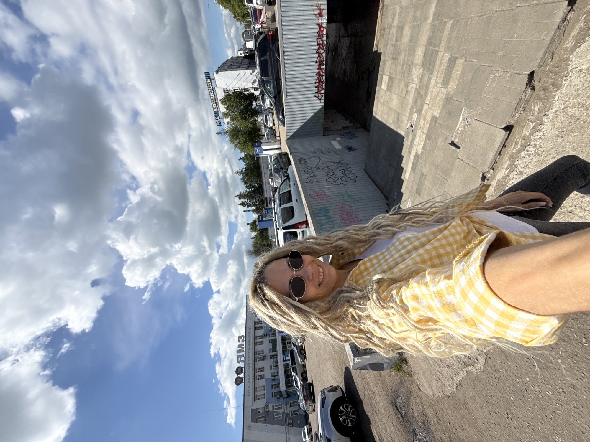 The image captures a selfie of a smiling young woman with long, blonde dreadlocks or very curly hair. She wears dark, round sunglasses and a yellow and white checkered shirt over a white top. Her arm is visible as she holds the camera, positioned against a backdrop of an urban industrial setting.
The scene unfolds on a bright, sunny day, with a clear blue sky dotted by fluffy white clouds. In the background, an expansive paved parking lot is filled with numerous cars and vans. Several buildings are visible, including a prominent white industrial building featuring a large sign in Cyrillic text that reads "ЯРОСЛАВСКИЙ МОТОРНЫЙ ЗАВОД" (Yaroslavl Motor Plant), with a smaller "ЯМЗ" (YaMZ) logo underneath.
To the woman's right, a corrugated metal structure, possibly part of an underpass or utility enclosure, is covered in graffiti. The woman appears to be standing on an elevated concrete ledge or wall, looking down at the parking area and buildings. Her cheerful smile suggests a positive mood.