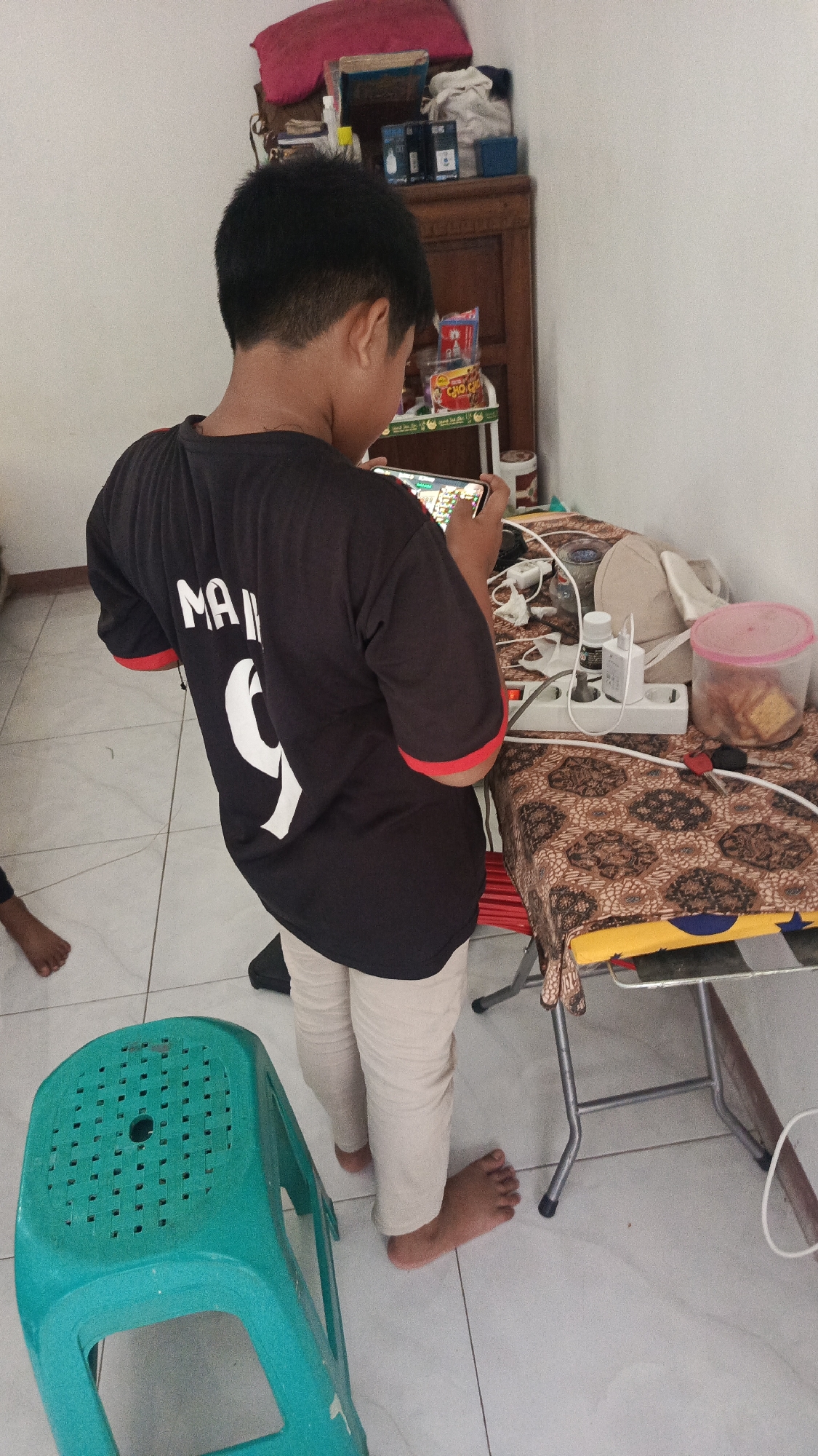A young boy, likely Indonesian, stands in a room and plays on a mobile phone. He wears a black t-shirt with red trim and light-colored pants, and his feet are bare. The room appears to be a home in Parung Panjang, Indonesia, based on the context provided. The boy is positioned facing away from the camera, engrossed in his phone.
In the background, a wooden cabinet holds various items, including what appears to be toiletries and snacks. A table covered with a patterned cloth is to the boy's right, cluttered with electrical items such as an extension cord and a power strip with plugs. A pink plastic container with food items sits on the table. Next to the boy on the left is a bright green plastic stool. The floor is tiled in a light color, and the walls are painted white. The lighting suggests an indoor setting, possibly during the daytime.