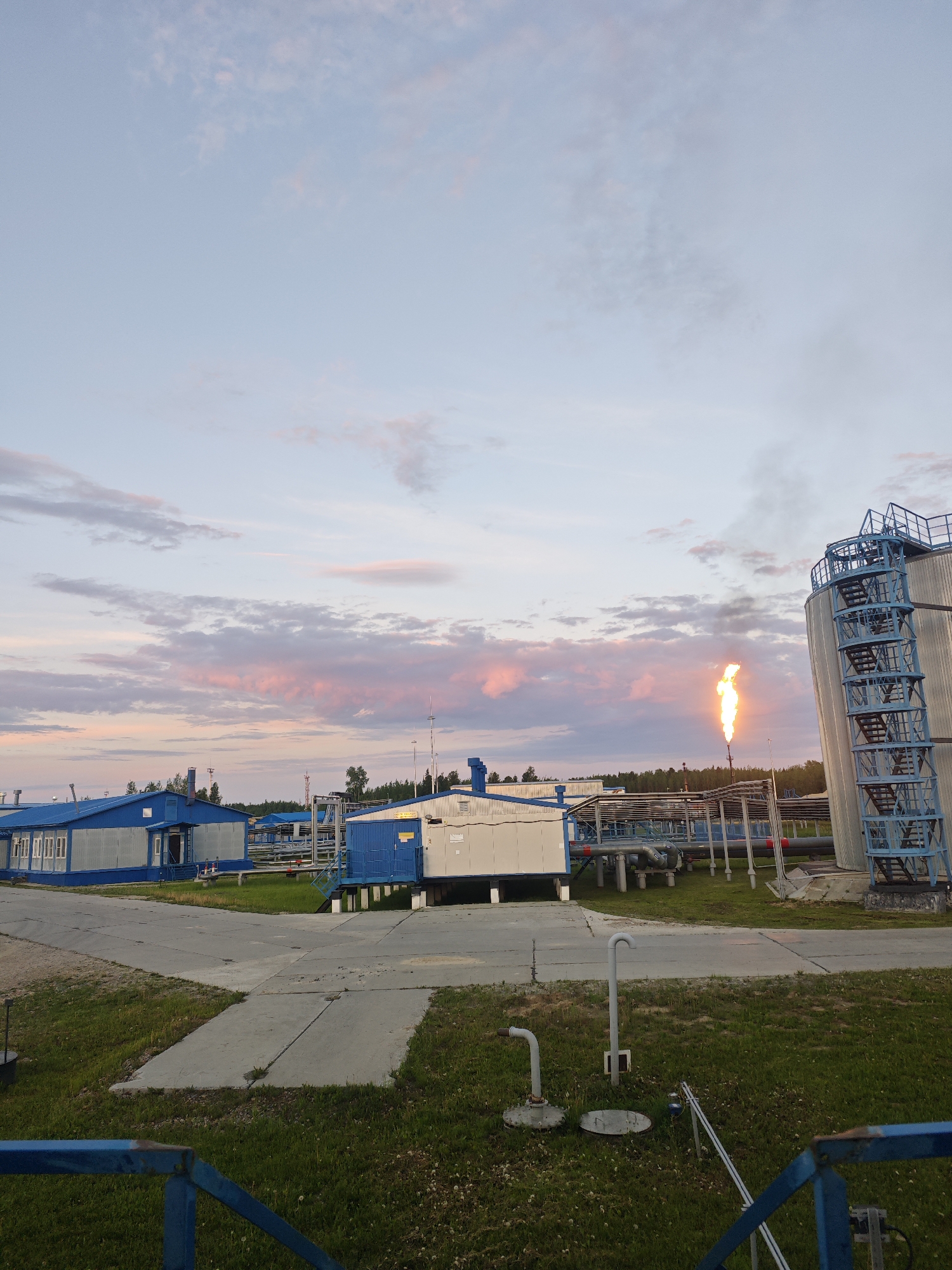 An industrial complex is depicted at dusk. The sky is a soft blue with streaks of pink clouds, suggesting either sunrise or sunset. In the foreground, a blue metal railing partially obstructs the view. Beyond the railing, a concrete pathway winds through a grassy area dotted with small white flowers.
Several prefabricated buildings, painted in shades of blue and white, are situated in the middle ground. These buildings appear to be modular units, possibly administrative offices or equipment shelters. Various industrial pipes and structures, some elevated on concrete supports, are visible behind and between the buildings.
To the right of the frame, a tall, cylindrical metal storage tank with a blue external staircase dominates the scene. In front of this tank and the industrial structures, a flare stack is actively burning, emitting a bright orange flame and a plume of smoke that rises into the sky. The overall impression is of an operational industrial facility at the close of day.