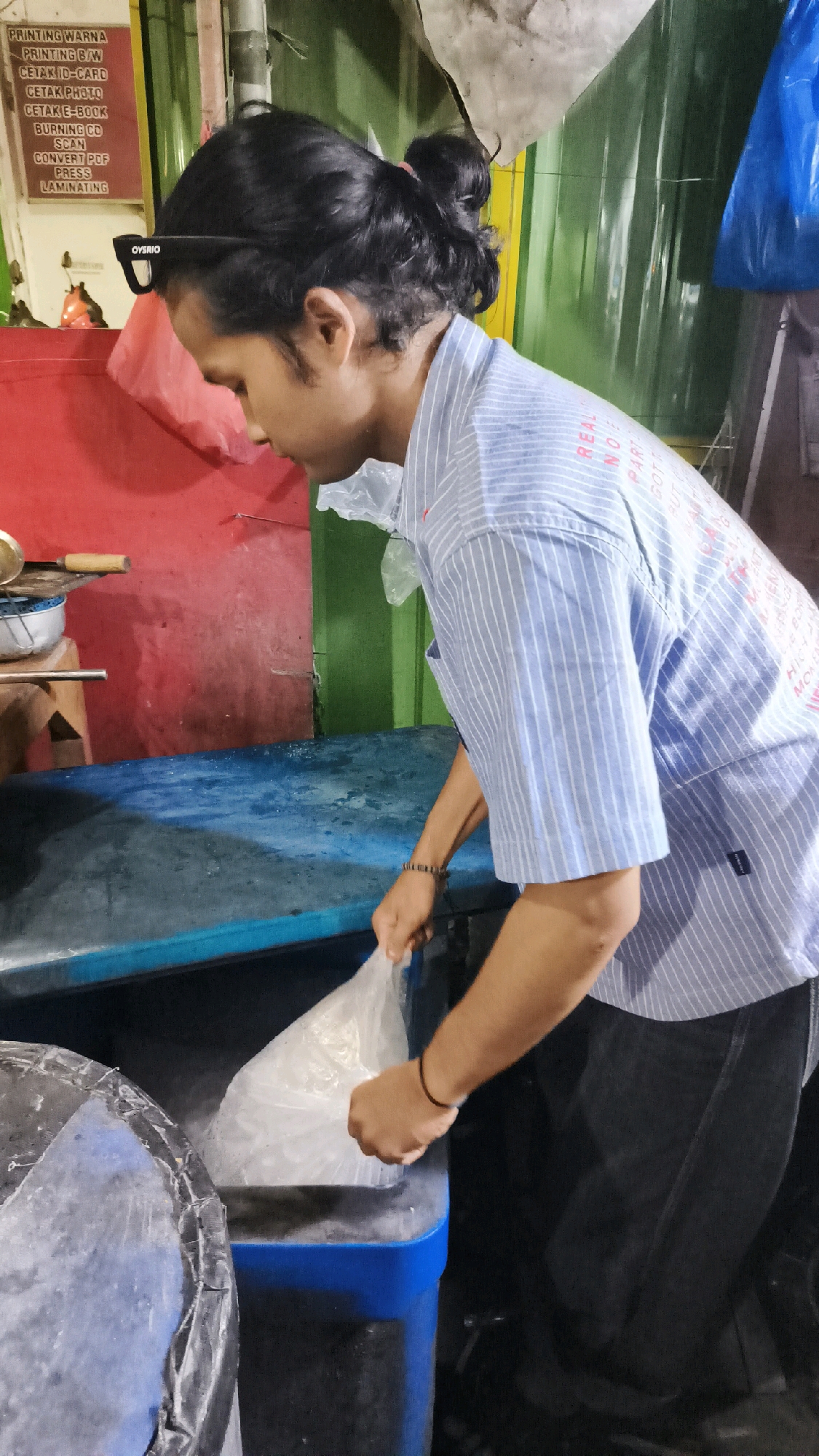 The image shows a person in a blue and white striped shirt, with their hair pulled back in a bun, wearing black-rimmed glasses. The person is holding a clear plastic bag filled with what appears to be ice and is in the process of placing it into a dark blue bin. The background features a red wall on the left, a green surface on the right, and a sign with Indonesian text that appears to list printing services. The scene suggests an outdoor or semi-outdoor setting, possibly a food stall or service area, located in Kartasura, Indonesia.