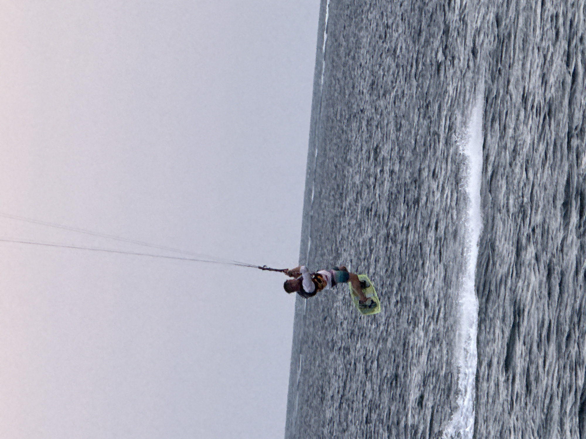 A kiteboarder is captured mid-air, suspended above choppy, dark blue ocean water under a pale, overcast sky. The kiteboarder, wearing a white long-sleeved shirt, a black and white vest, and multi-colored board shorts, is holding onto a kite control bar attached to a kite by thin lines, which are visible against the sky. The kiteboarder is positioned to the left of the frame, with their legs bent and feet strapped into a light green kiteboard. A spray of white water is visible behind the board, indicating recent movement through the water. The horizon line where the ocean meets the sky is slightly blurred. The overall scene is indicative of a watersport activity. The location is identified as Denpasar, Indonesia.