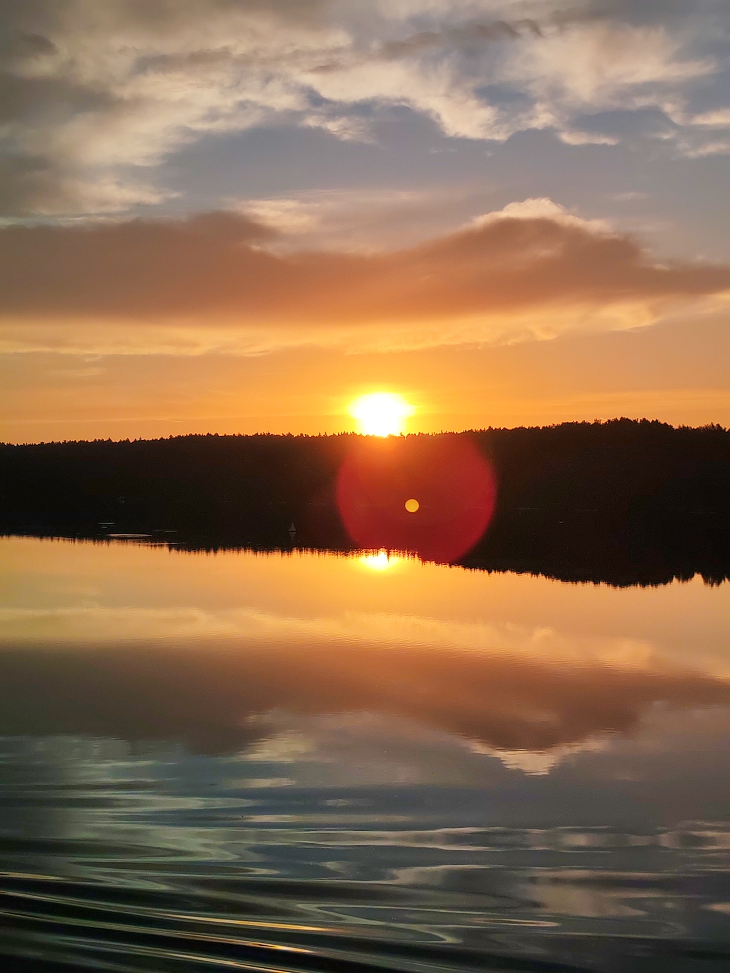 A vertical, wide shot captures a sunset over a body of water, likely a lake or slow-moving river, near the village of Terekhovo, Russia. The sun, a bright white disc with a distinct red lens flare to its right, is partially obscured by a dark, treeline-covered ridge that bisects the image horizontally. The sky above is a gradient of warm oranges and yellows near the horizon, transitioning to muted blues and grays with wispy clouds higher up. The water in the foreground reflects the sky and the silhouetted treeline, creating a mirrored image. Ripples on the water's surface, particularly in the bottom portion of the frame, suggest gentle movement or boat wake. The overall scene is serene and natural, with no visible human presence or activity beyond the implied wake in the water.