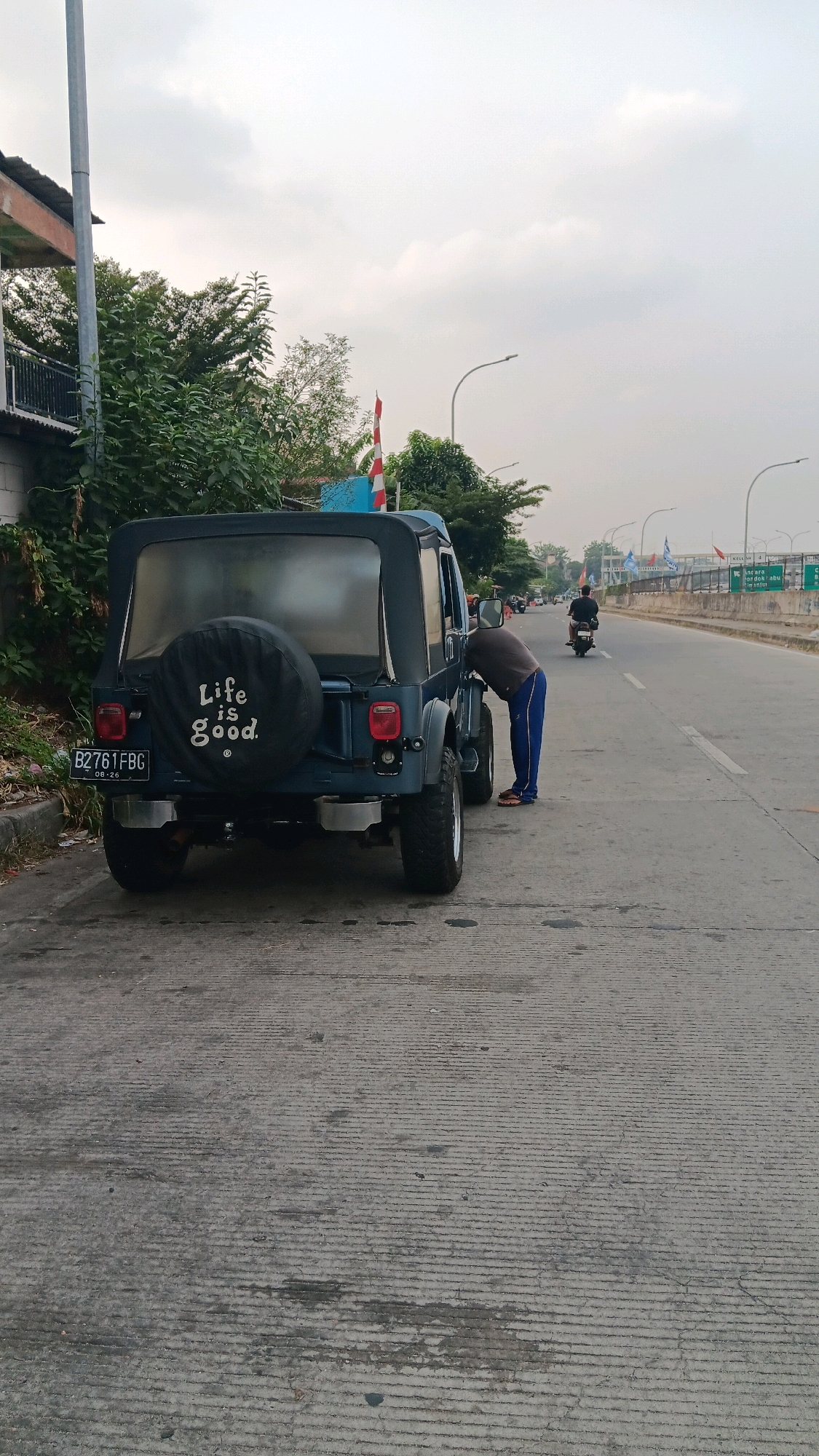 A dark blue jeep is parked on the side of a road in Depok, Indonesia. The jeep has a spare tire cover with the words "Life is good." written on it. A person is bending over the jeep, appearing to be inspecting it. The road is paved and there is a sidewalk on the left side of the image. Trees and buildings can be seen in the background. The sky is cloudy. A motorcycle is riding on the road in the distance.