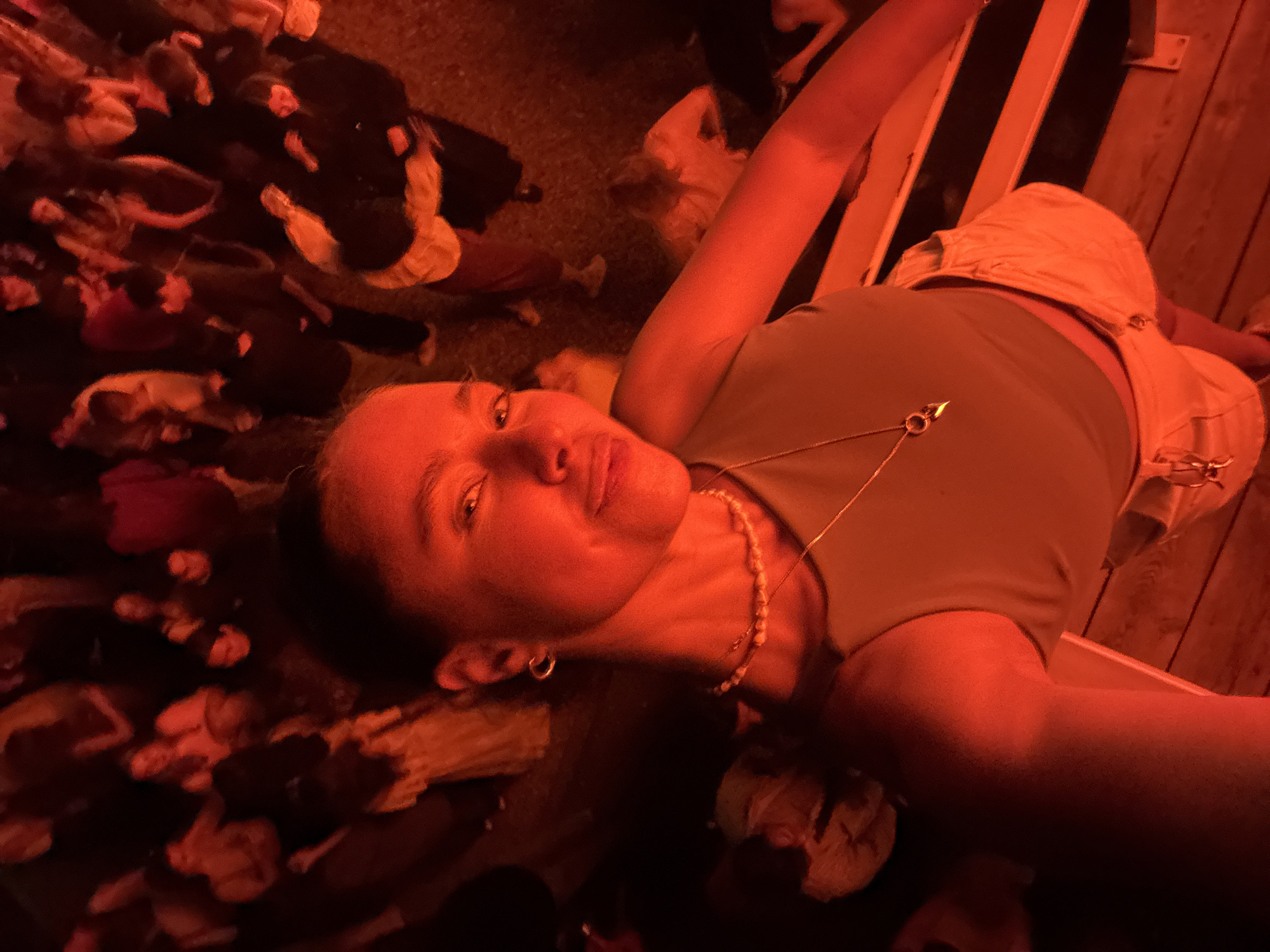A selfie-style photograph taken from a low angle depicts a person with fair skin, light brown hair pulled back, wearing a green tank top, beige shorts, and a gold necklace. The person is lying down on a wooden surface with a railing visible in the background. The environment appears to be crowded, with numerous people visible in the background, possibly at an outdoor event or gathering, illuminated by red light. No information connects the scene to Kazan, Russia.