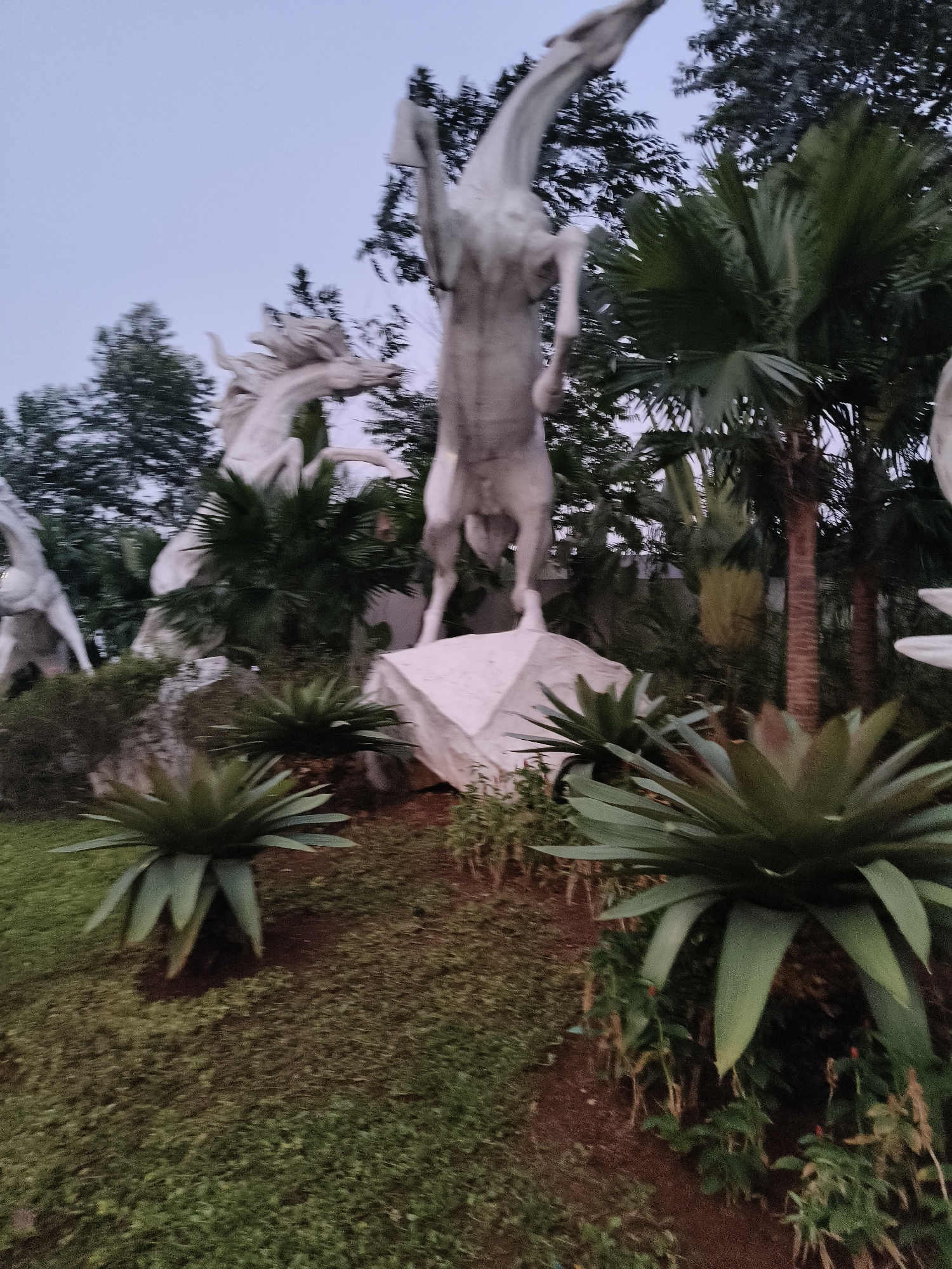 The image depicts a surreal outdoor art installation featuring multiple large, white sculptures of rearing horses against a backdrop of lush tropical foliage and trees. The sky is overcast and appears to be during twilight or early morning. In the foreground, there are spiky green plants, possibly bromeliads or agaves, and a grassy area. The sculptures are in various dynamic poses, suggesting movement and power. The overall impression is one of a fantastical or mythical setting. This installation is located at the De Maja House, a historical and cultural site in Cisauk, South Tangerang, Banten, Indonesia.