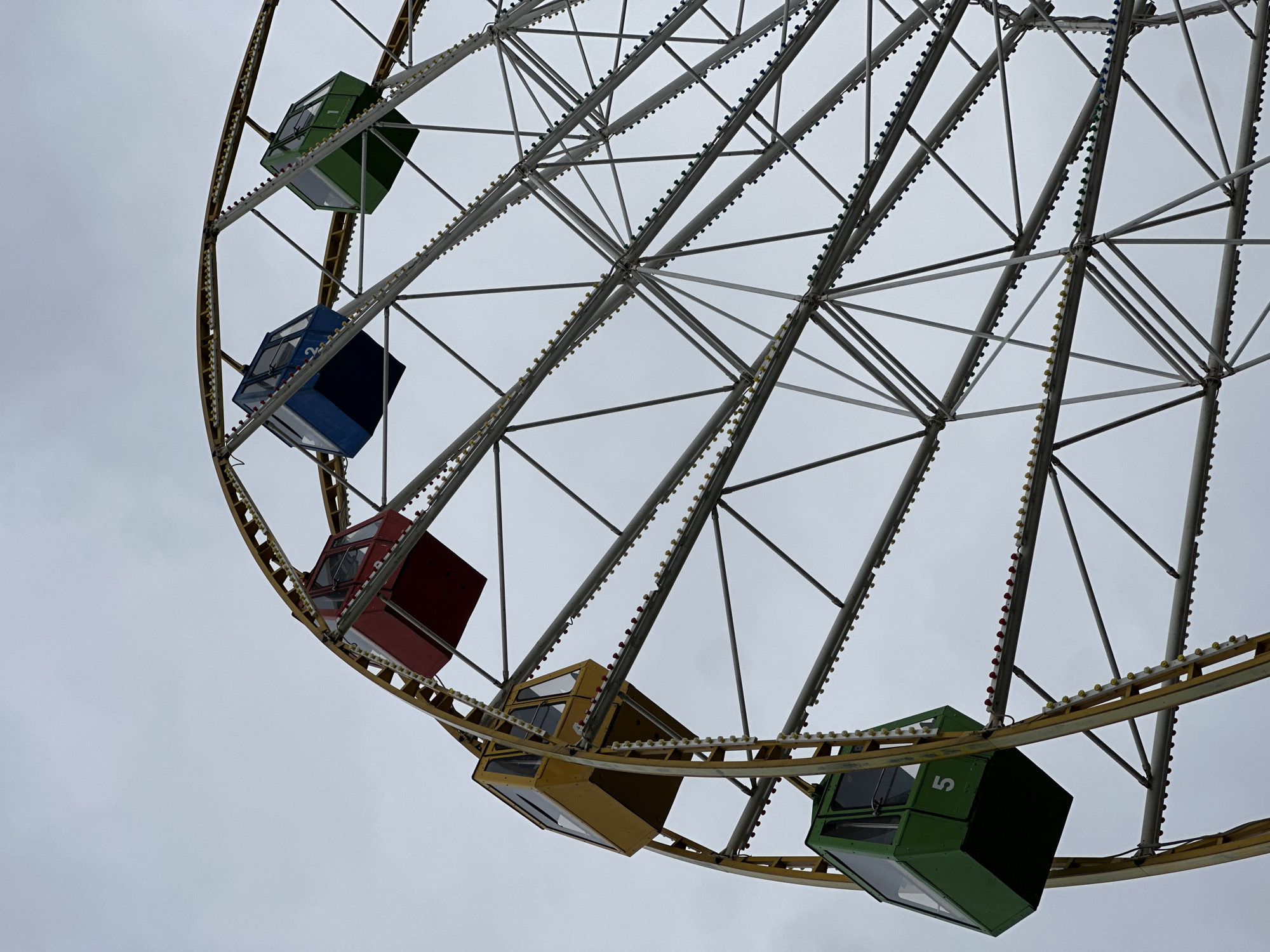 The image depicts a partial view of a Ferris wheel against a gray sky. The Ferris wheel has a metallic structure, and several colored cabins are visible: green, blue, red, and yellow. The cabins appear to be unoccupied. The cabins are attached to the perimeter of the Ferris wheel. The photo is taken from a low angle, emphasizing the height and structure of the Ferris wheel. Based on image search results, this may be located in Stavropol, Russia.