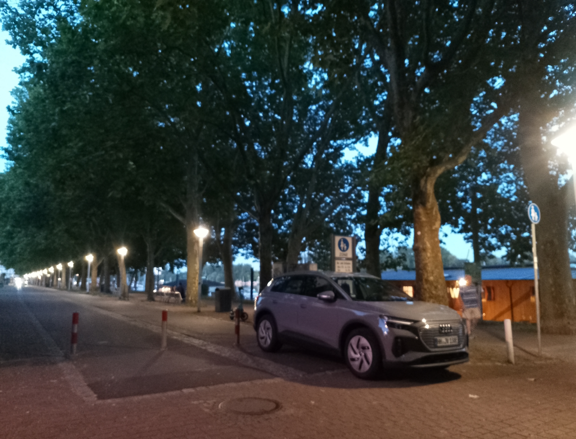 The image shows a street lined with trees and lampposts, with a light-colored SUV parked on the right side of the road. The sky is dark blue, suggesting it is evening or early morning. The road appears to be paved with asphalt and cobblestones. There are some bollards on the side of the road. In the background, there are buildings with illuminated windows and a few people are visible, one standing near the car. There is a traffic sign and a garbage bin near the parked car. The scene depicts a public street with parked vehicles and pedestrian access.