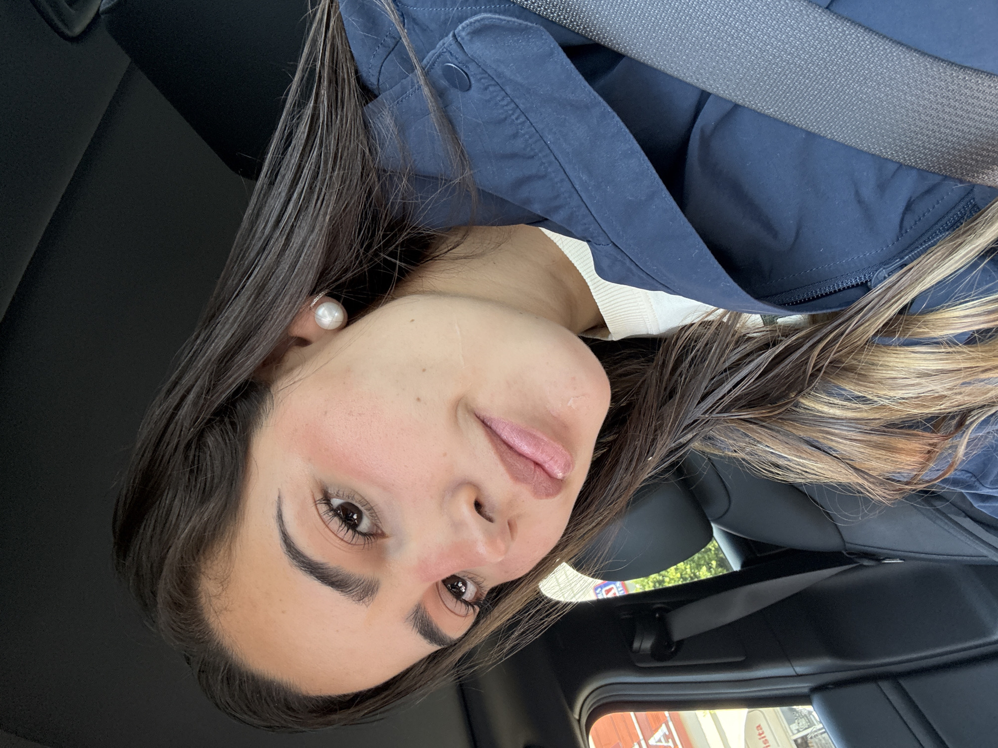A woman is wearing a pearl earring, a white sweater, and a navy blue jacket with a seatbelt buckled around her torso. She is in a car, potentially driving in Puebla, Mexico.