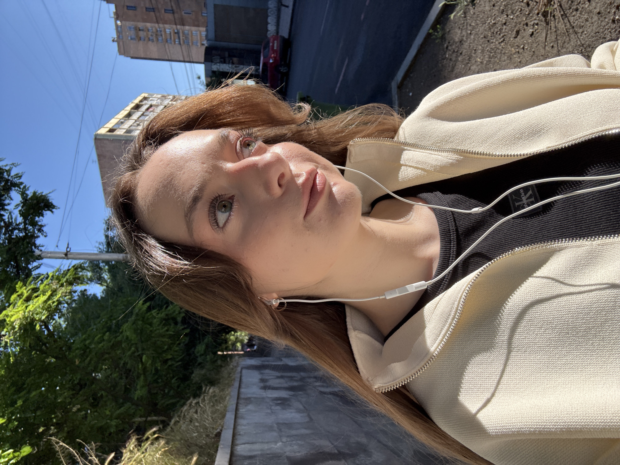 A young woman with fair skin and light brown hair is taking a selfie outdoors on a sunny day. She is wearing a black top and a cream-colored zip-up jacket. White earphones are visible, inserted in her ears. The background includes a street, a building with multiple stories, trees, and a blue sky. The photo was taken in the city of Yerevan, Armenia.