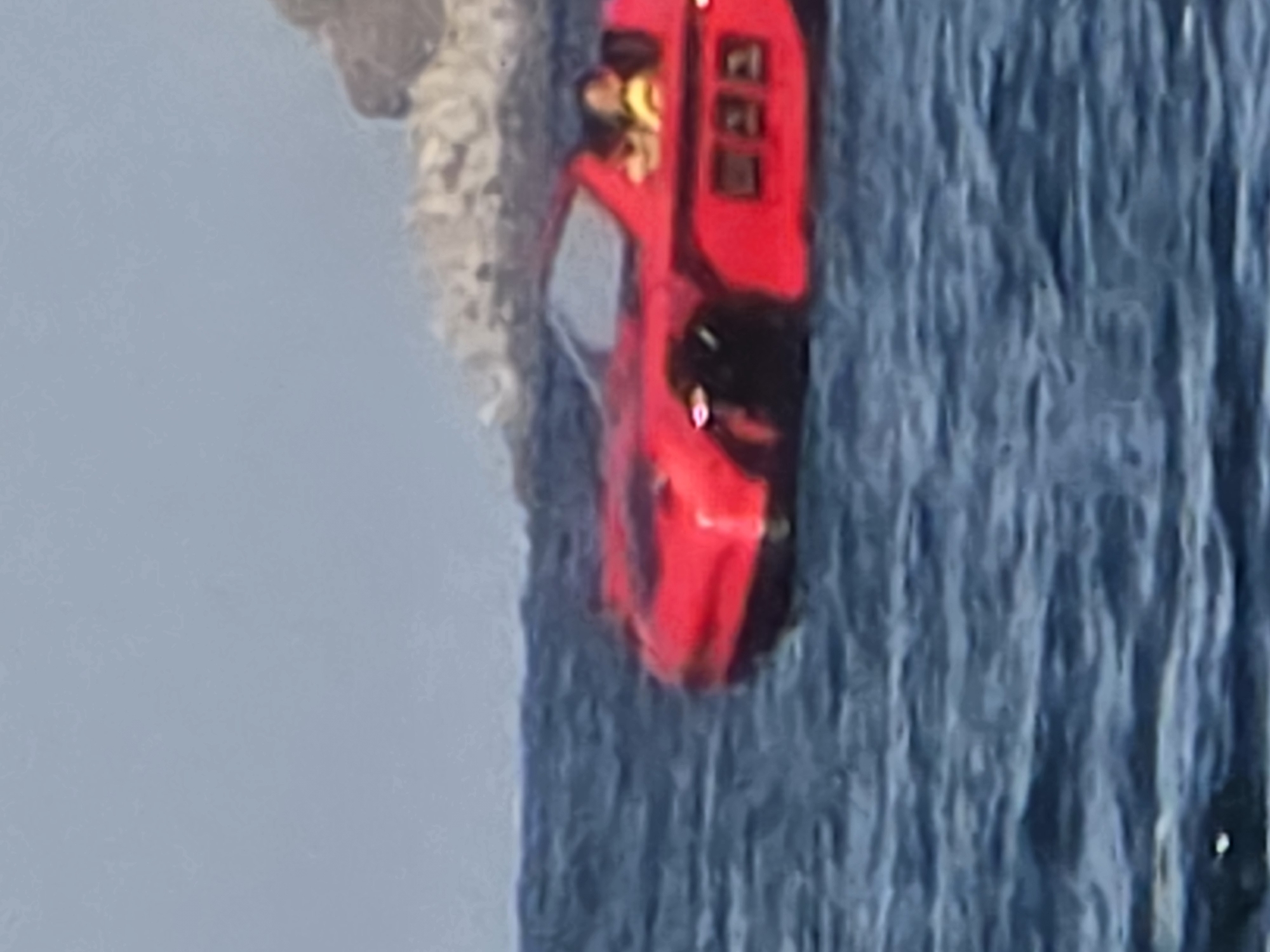 A red amphibious car, designed to resemble a convertible sports car, is navigating through choppy blue water. Two individuals are visible inside the vehicle; one appears to be a person with short, dark hair wearing a yellow top, and the other is partially obscured. The amphibious car has a sleek, low-profile design with visible square panels on its side. In the background, a rocky, hilly coastline is visible under a pale blue sky. The scene takes place in the sea near Chernomorets, Bulgaria.