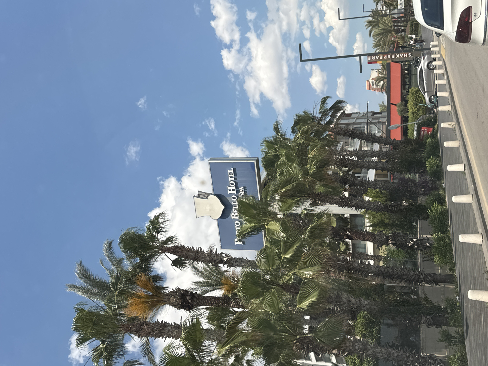 The image shows a street scene in Konyaaltı, Turkey, featuring the Porto Bello Hotel & Spa. A row of palm trees lines the street, partially obscuring the hotel's sign. In the background, a building with a "Shakespeare" sign is visible. A white car is parked along the roadside next to concrete bollards. The sky is blue with scattered white clouds.