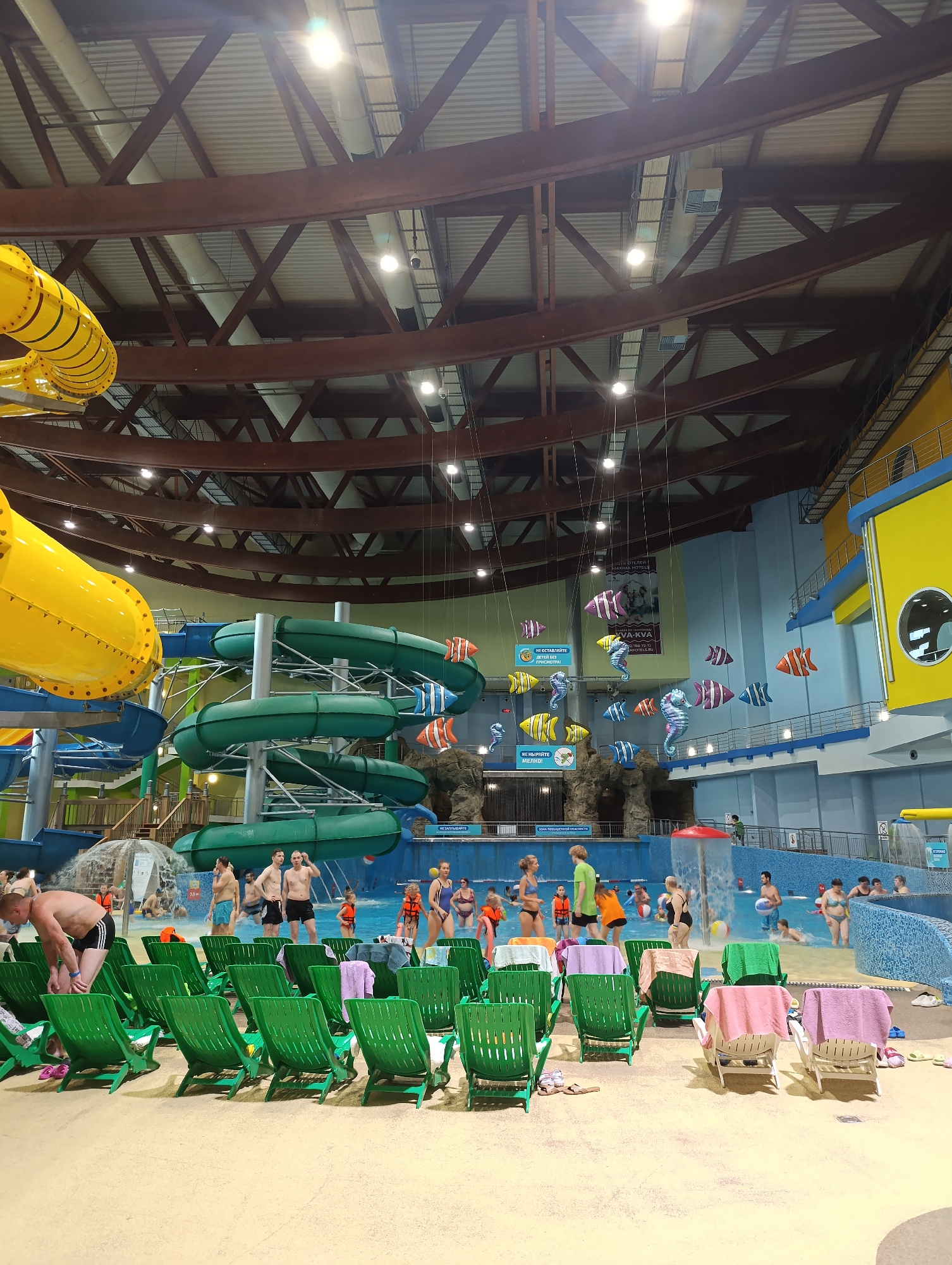 The image shows an indoor water park. In the foreground, there are rows of green lounge chairs with towels draped over them. In the midground, there is a large pool area with several people swimming and playing. There are also water slides in the background, with a large yellow slide on the left and green slides in the center. The ceiling of the water park is high with exposed beams and lighting fixtures. Decorations resembling fish and seahorses are suspended from the ceiling. The overall environment appears to be bright and recreational.