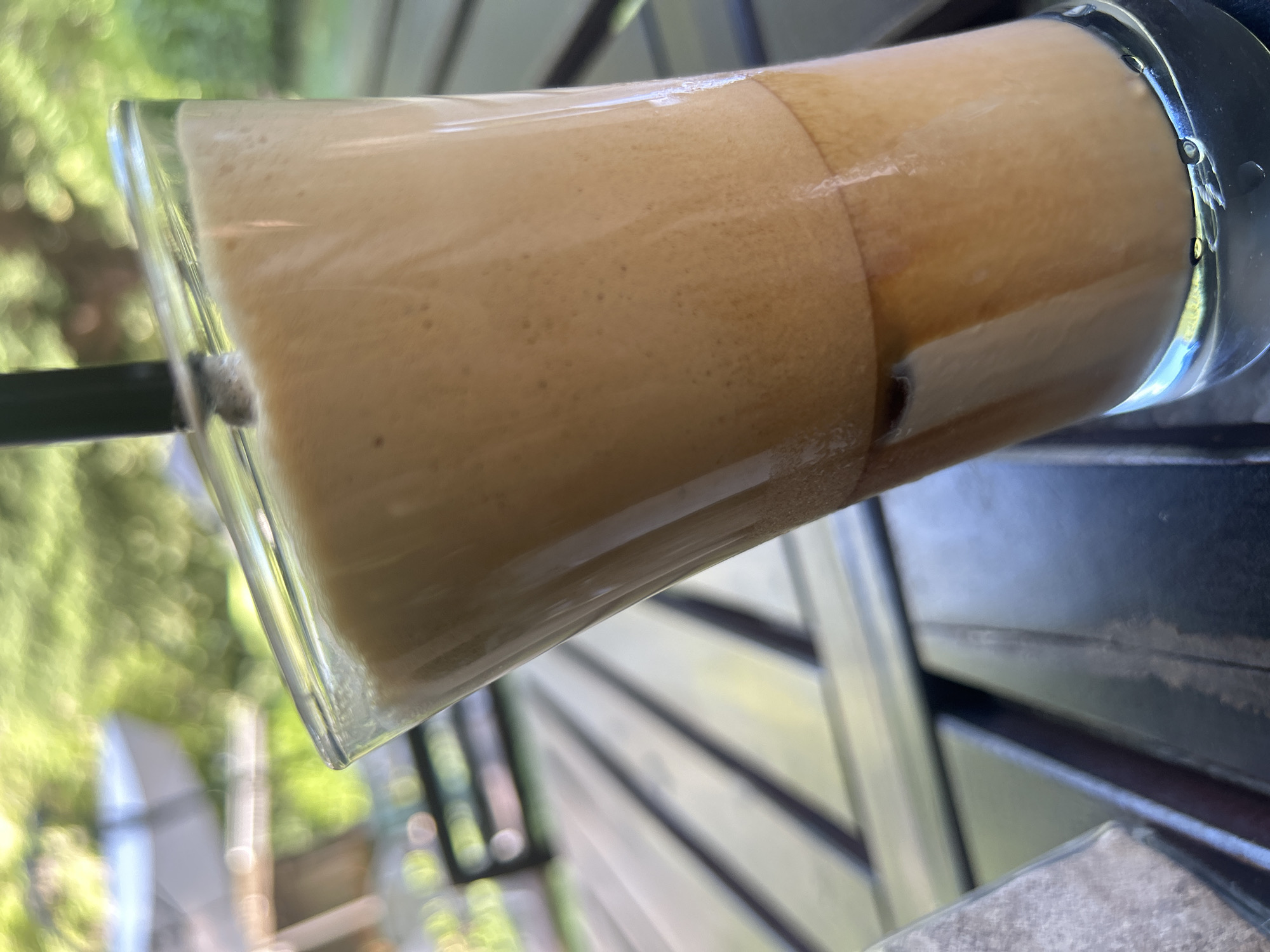 A tall, clear glass of iced coffee with a frothy top and a dark liquid layer at the bottom sits on a dark wooden table. A black straw is inserted into the drink. The table appears to be outdoors, with blurred green foliage and sunlight in the background, suggesting a cafe or restaurant setting in Varna, Bulgaria. The focus is on the beverage, with the surroundings softly out of focus.