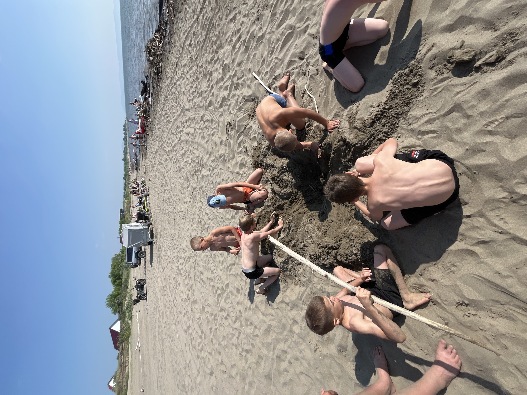 In Krasnoturansk, Russia, on a sunny day, a group of young boys are playing in the sand near the water. They are digging a hole with sticks and their hands. In the background, there are trees and buildings, and the water stretches out to the horizon.