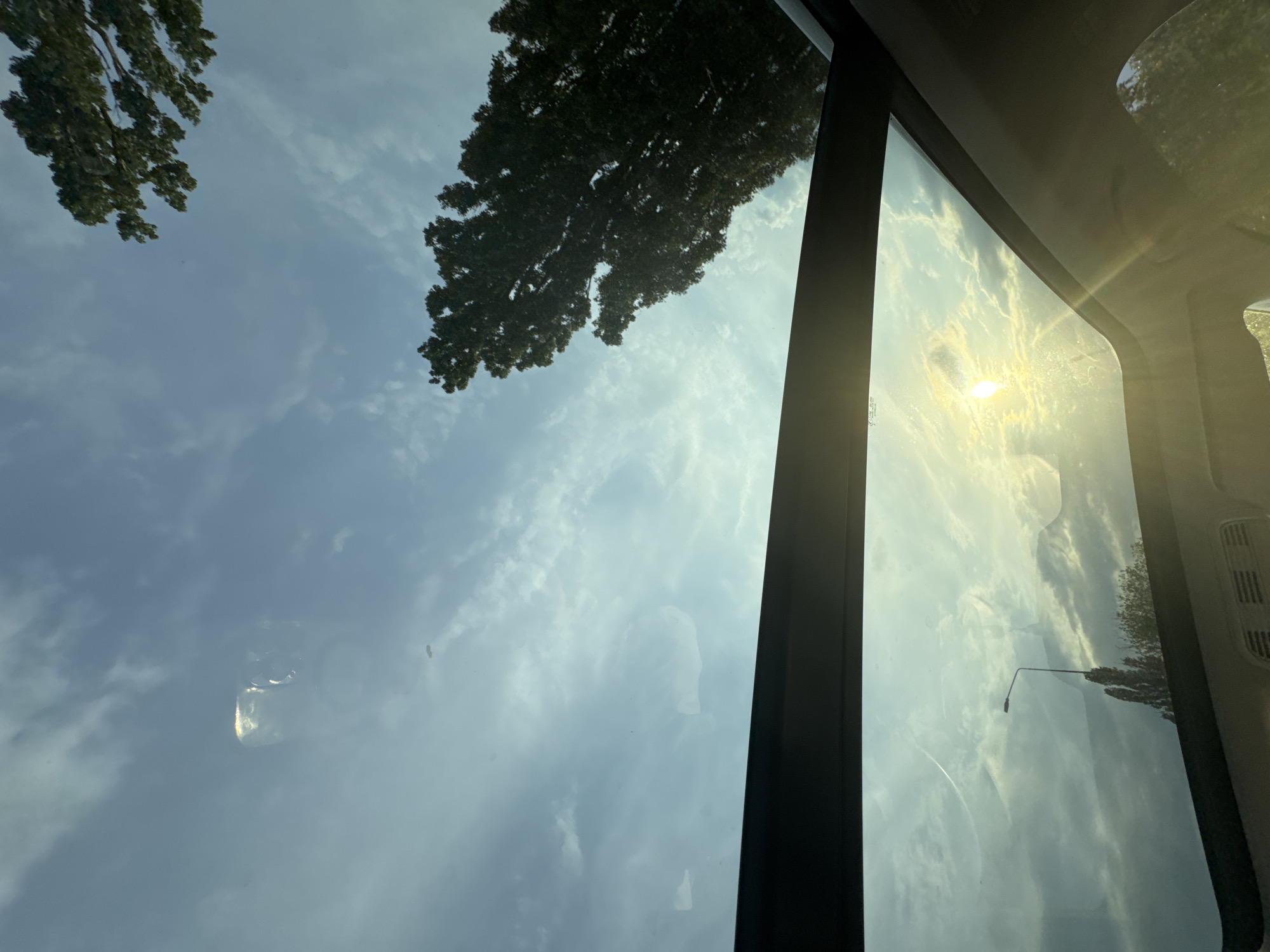The image, captured in Suprunivka, Ukraine, presents an upward view from inside a vehicle through its panoramic glass sunroof. The sky displays a light blue color with scattered white, wispy clouds. The sun, positioned low in the sky, manifests as a bright, circular flare in the lower right quadrant of the sunroof, casting a golden-yellow light across portions of the clouds and the glass surface. The dark green foliage of two large trees is prominently visible against the sky in the upper right and upper left areas of the frame. A distinct reflection of an interior vehicle light fixture, appearing as a rectangular shape with a bright outline, is observed on the upper left section of the sunroof glass. In the lower left, below the sunroof's framing, a dark street light pole with a single luminaire extends vertically, accompanied by the upper crown of another deciduous tree. The black frame of the sunroof and sections of the vehicle's gray interior headliner delineate the boundaries of the scene. No individuals or discernible actions are depicted.