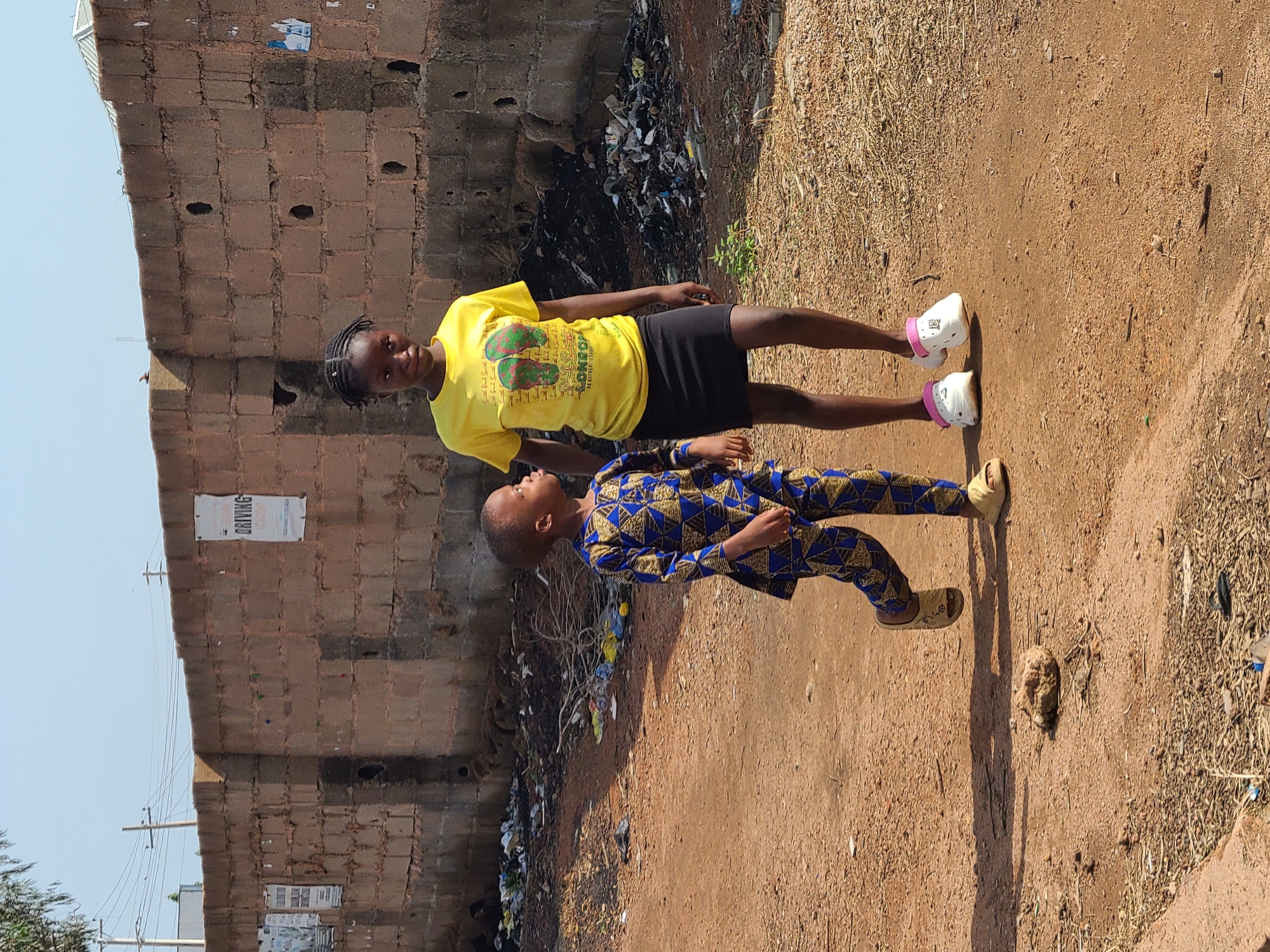 Two young individuals, a girl and a boy, are walking along a dirt road in Gura Top, Nigeria. The girl, older of the two, wears a bright yellow t-shirt with a graphic of two flip-flops and black lettering, paired with a black skirt and white clogs with pink accents. The boy is dressed in a blue and gold patterned matching set, including a long-sleeved shirt and pants, and wears light brown slip-on sandals. They are walking away from the camera, towards a brick wall that borders the road. The wall is constructed of rough, reddish-brown bricks and has several posters or signs attached to it, one of which appears to be for a "DRIVING ACADEMY." A considerable amount of debris and trash is visible along the base of the wall and on the ground, indicating a somewhat neglected environment. Power lines are visible against a clear blue sky. The overall scene depicts a moment of everyday life in an urban or semi-urban setting in Nigeria.