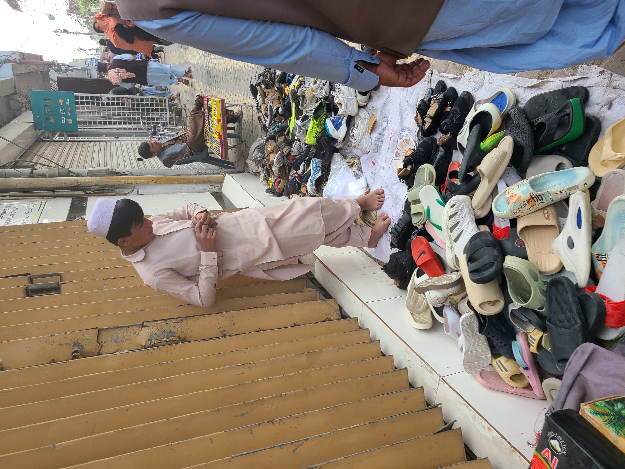 The image depicts a street scene in Quetta City Tehsil, Pakistan. A young boy in a light pink shalwar kameez and white cap is lying on a yellow metal roll-up storefront shutter. Adjacent to him, various types of footwear, mostly slippers and sandals, are piled on a white sheet or tarp. The scene includes other people in the background, an ATM sign, and parts of buildings indicative of an urban setting.