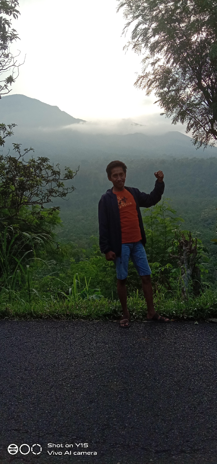 The image depicts an adult male standing on a dark gray asphalt road overlooking a vast, lush green mountainous landscape in Ranuagung, Indonesia. The man, of apparent Southeast Asian descent, is positioned slightly right of center, facing the camera with a subtle smile. He wears a dark blue unzipped hooded jacket over an orange t-shirt, light blue denim shorts, and dark sandals. His left arm is bent at the elbow, with his fist clenched and raised above his left shoulder. The background features successive layers of forested hills and mountains, with distant peaks partially obscured by atmospheric mist or haze under a bright, overcast sky. Green grass and foliage line the edge of the asphalt road in the foreground.