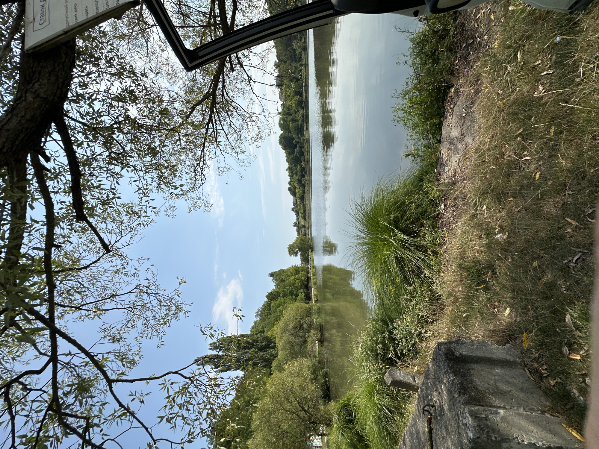 The image depicts a serene lake scene in Makiv, Ukraine, on a clear, sunny day. The foreground features a concrete structure on the left, partially covered by dry grass and fallen leaves. To the right of the concrete, tall green grass and smaller vegetation grow. A black car door is open on the far right of the frame, with its interior partially visible.
The middle ground is dominated by the calm surface of the lake, reflecting the blue sky, scattered white clouds, and the lush green trees that line the opposite shore. These trees vary in height and density, creating a natural backdrop. In the distance, a few houses are visible nestled among the trees on the far bank, suggesting the presence of a small settlement.
The sky above is a bright blue with a few wispy white clouds. Branches and leaves from a large tree hang into the frame from the top, framing the view of the lake and sky. A sign is attached to the tree on the right, partially obscured by branches and the car door. The overall impression is one of tranquility and natural beauty.