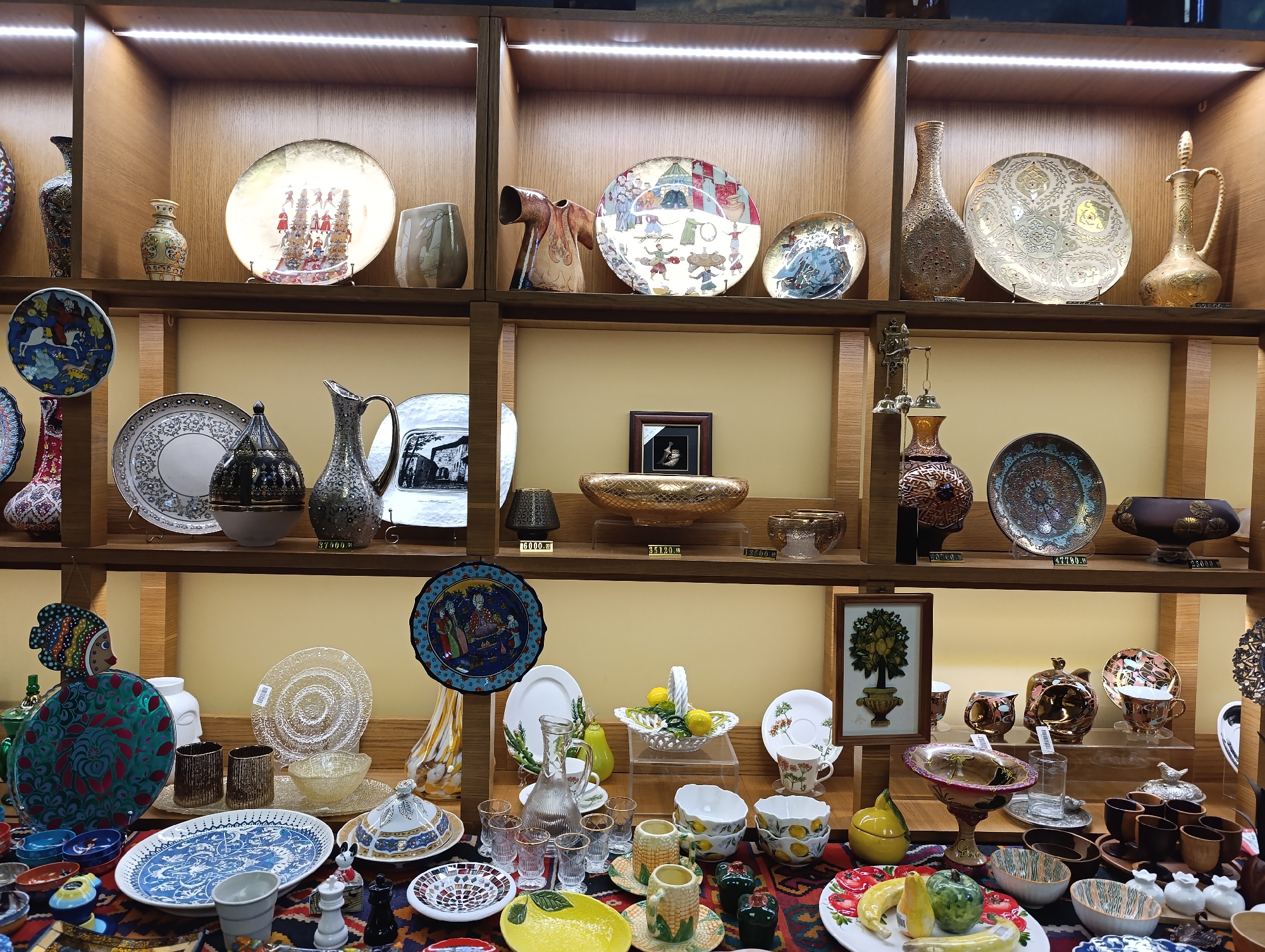 The image depicts a display of decorative ceramic and glassware items on wooden shelves in a retail setting. The items include plates, vases, bowls, and pitchers, many with intricate patterns and metallic finishes. The scene appears to be indoors, with lighting illuminating the shelves. The items are arranged with care, suggesting a curated collection for sale. There are no people visible in the image, and no action is taking place. The overall impression is of a well-stocked shop specializing in artistic homeware. The presence of price tags suggests that this is a commercial establishment. The specific location in Moscow, Russia, is not indicated by any visible elements in the image.