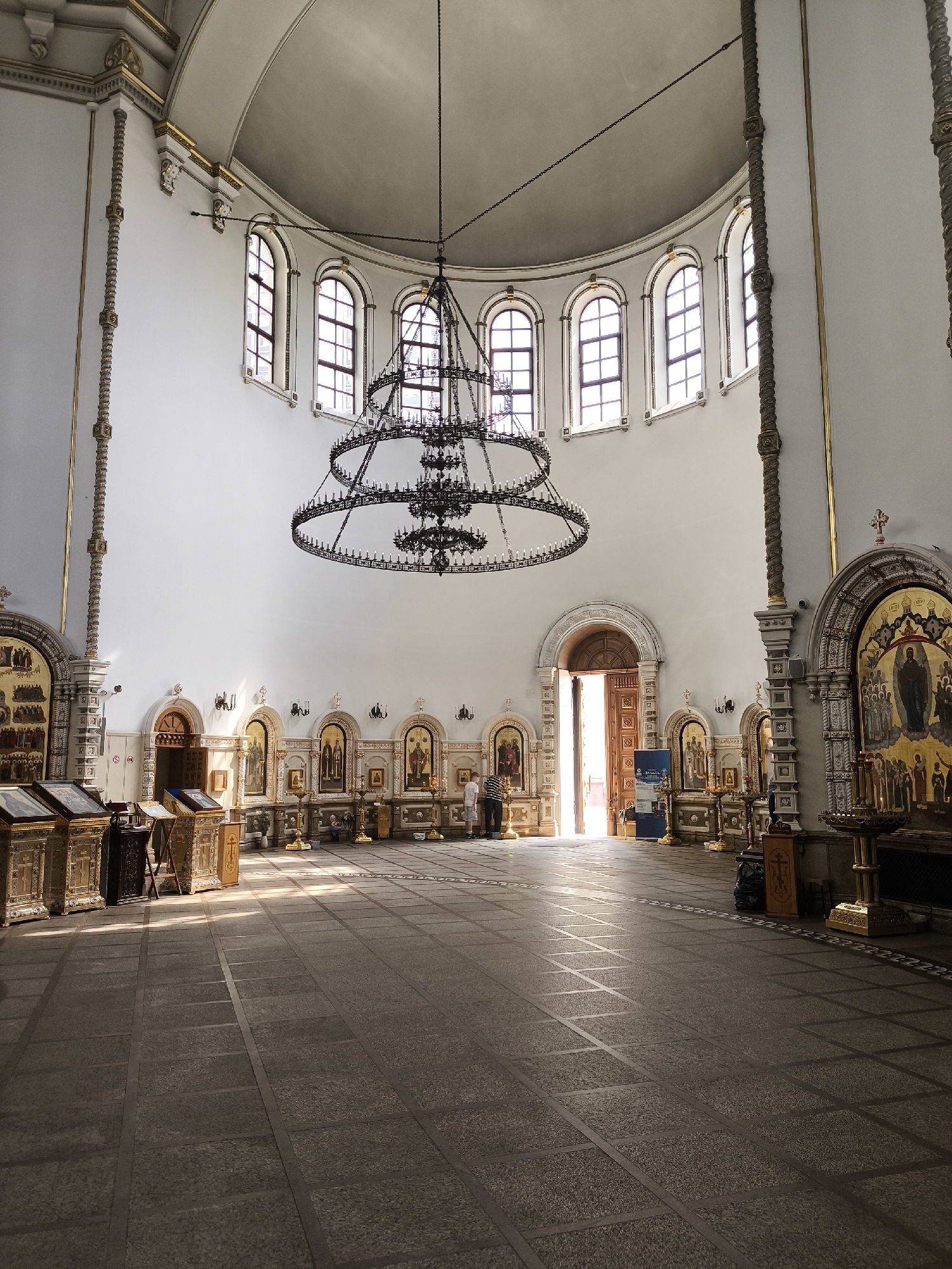 The image depicts the interior of a grand, ornate church. Sunlight streams through arched windows on the upper wall, illuminating a vast, tiled floor. A large, multi-tiered chandelier hangs prominently from the domed ceiling. Along the walls are numerous icons displayed in ornate frames, with several people standing in the space. To the left, what appear to be gilded prayer stands or podiums are positioned. In the center, an open doorway casts a bright light into the hall, revealing a glimpse of the outside. The walls and ceiling are painted in a light color, adorned with decorative molding and gold accents. Two individuals are visible near the entrance, and at least two more are further into the church, one standing by an icon and another near the open doorway. The overall impression is one of solemnity and architectural splendor.