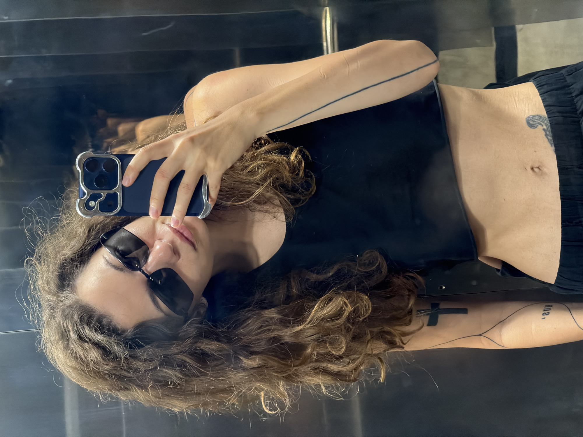 A person is taking a selfie in what appears to be a reflective surface, likely a mirror. They are wearing sunglasses and a black top, and their hair is wavy. The person is holding a smartphone in their right hand. Tattoos are visible on their arms and torso. The reflection suggests the environment might be indoors. The location in Đà Nẵng, Vietnam is not evident from the image.