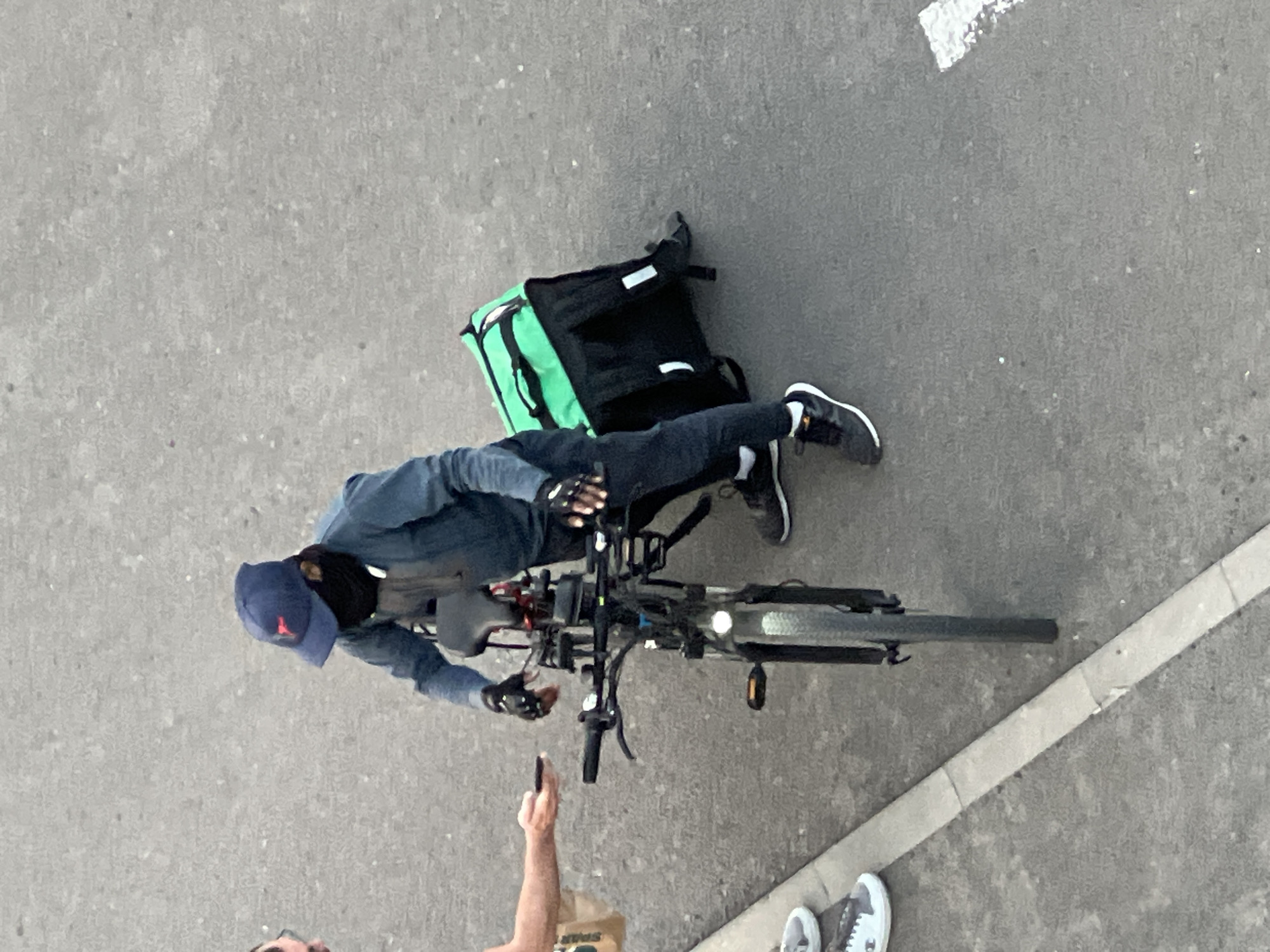 The image captures a scene on an asphalt surface, possibly a street or sidewalk, in Florești, Romania. In the center, a person on a bicycle is interacting with another individual. The person on the bicycle wears a blue baseball cap, a dark mask covering the lower face, a long-sleeved blue shirt, dark pants, and black athletic shoes. They are wearing black gloves with white accents. Attached to the rear of the bicycle is a large, bright green and black insulated delivery backpack. The other individual, partially visible on the left, is holding out a mobile phone and a brown paper bag, suggesting an exchange or transaction. The environment is predominantly gray asphalt, with a concrete curb running along the bottom left of the frame. The lighting suggests it is daytime.