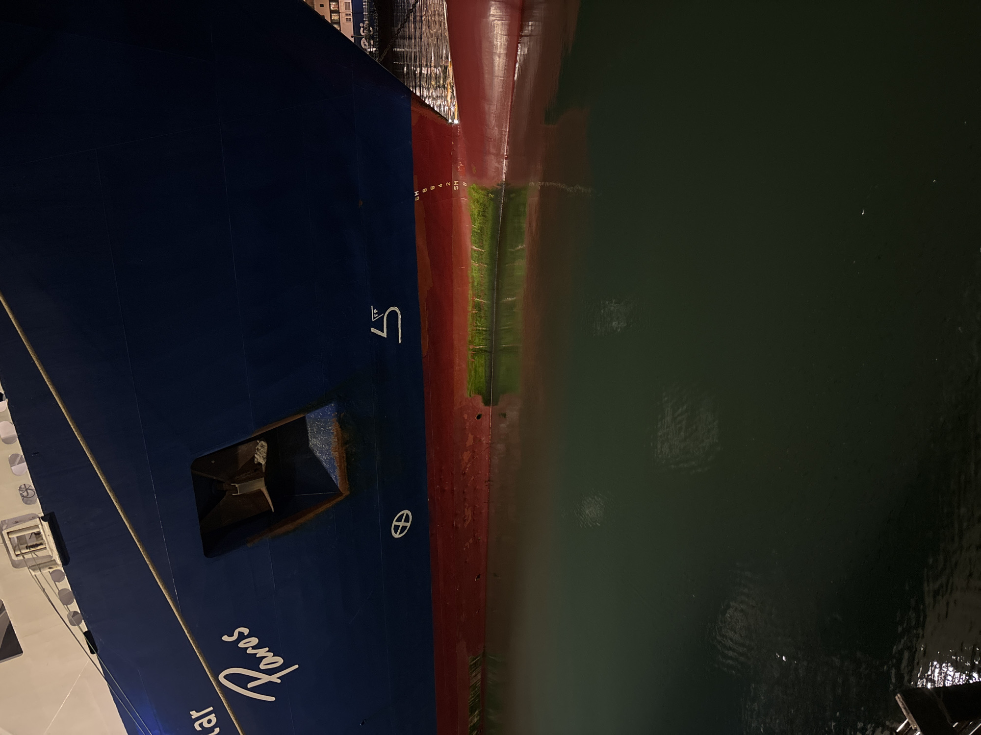 The image captures the bow of a ship, named "Star Faros", moored in the harbor of Piraeus, Greece. The ship's hull is painted with a color scheme of dark blue above the waterline and a red anti-fouling paint below. Algae growth is visible along the waterline. Depth markings are visible on the hull. The anchor housing is open, revealing the anchor. The water reflects the ship and some distant lights. The time of day is likely evening or night due to the lighting conditions.