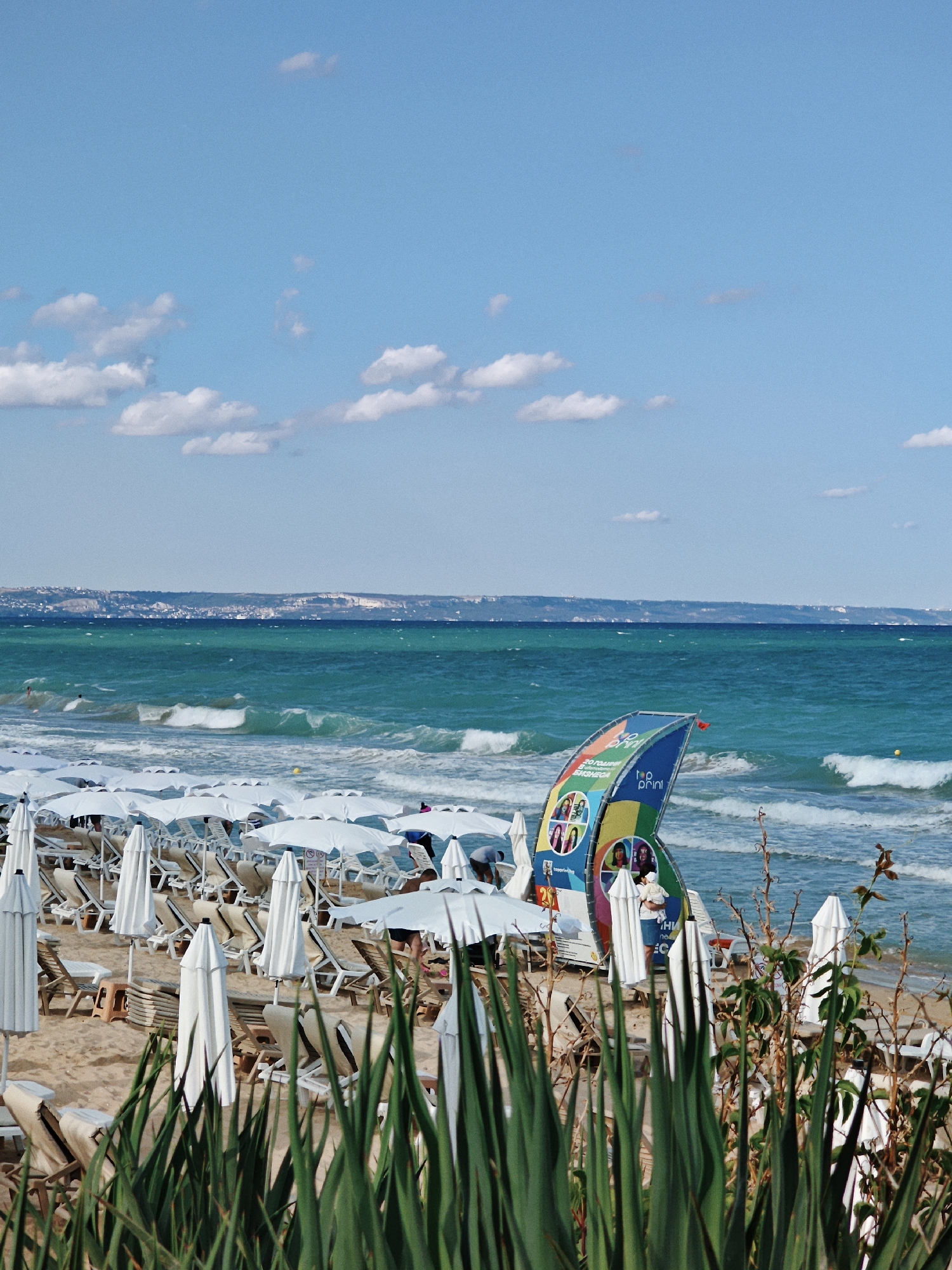 The image depicts a crowded beach scene with rows of white umbrellas and lounge chairs on the sand. The turquoise ocean stretches to the horizon, with waves breaking on the shore. In the foreground, tall green grass partially obscures the view of the beach. A colorful banner with images of people stands on the beach near the water. In the distance, a city or town is visible along the coastline. The sky is blue with scattered white clouds. The overall impression is a busy summer day at the beach.