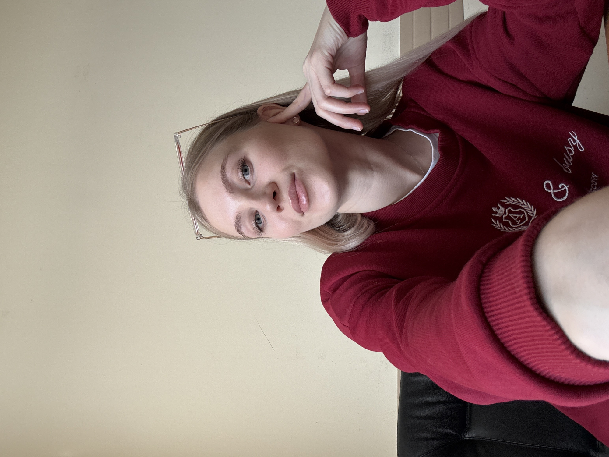 A person, presumably female, is captured in a close-up, angled selfie. The person is wearing glasses and a maroon sweatshirt with white lettering and a logo on the chest. Her blond hair is partially obscuring one side of her face. Her hand is near her ear, possibly adjusting her hair or glasses. The background consists of a light tan wall and a section of what appears to be a black leather chair. The image does not provide any information about the location in the city of Unknown, Unknown.