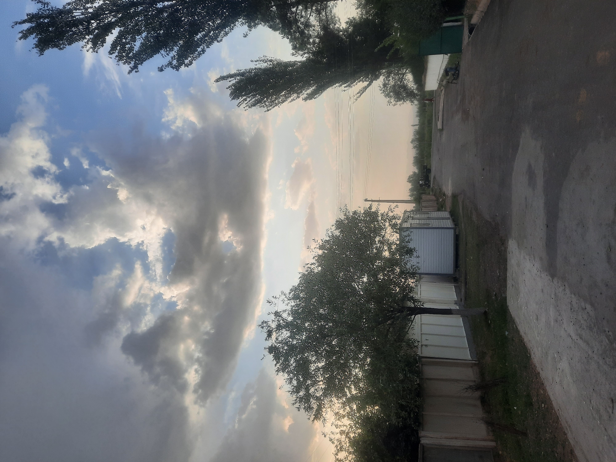 The image depicts an outdoor scene on a paved road in Voseaobad, Tajikistan. The sky is filled with dramatic, layered clouds, with patches of blue sky visible between them. On the left side of the road, a light-colored fence runs along the property line, with a small, bare-trunked tree in front of it. To the right of the tree are two modular white structures. Further down the road, tall, slender trees with dark foliage are prominent on the right side. The road appears to be an asphalt surface with some lighter patches, suggesting it might be wet in places. In the distance, there are more trees and vegetation, and mountains are faintly visible on the horizon. A utility pole with multiple wires is visible near the center-left of the image. The overall impression is that of a rural or semi-urban setting with a dramatic sky.