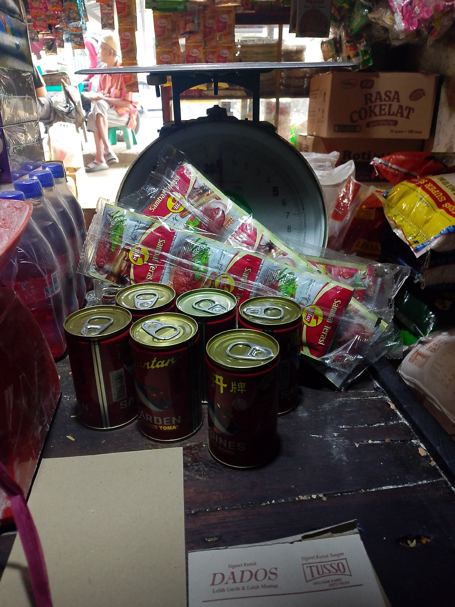 The image depicts a close-up view of a counter in a small, likely informal, store in Palembang, Indonesia. The counter is cluttered with various items. Prominently displayed are several red cans with gold pull-tabs, some of which have labels that partially read "SARDINES" and appear to be canned fish. To the left, several plastic bottles, possibly containing beverages, are visible. Above the cans, a clear plastic package holds multiple sachets of what appears to be a spicy condiment, labeled "Sambal Terasi." In the background, a large, old-fashioned mechanical scale with a round dial is situated on a wooden surface. The store environment is suggested by the presence of stacked boxes with text like "RASA COKELAT" (Chocolate Flavor) and other packaged goods hanging in the background, typical of a traditional Indonesian warung or kiosk. Natural light filters in from an opening in the background, illuminating the scene and casting shadows. The overall impression is of a busy, functional retail space.
