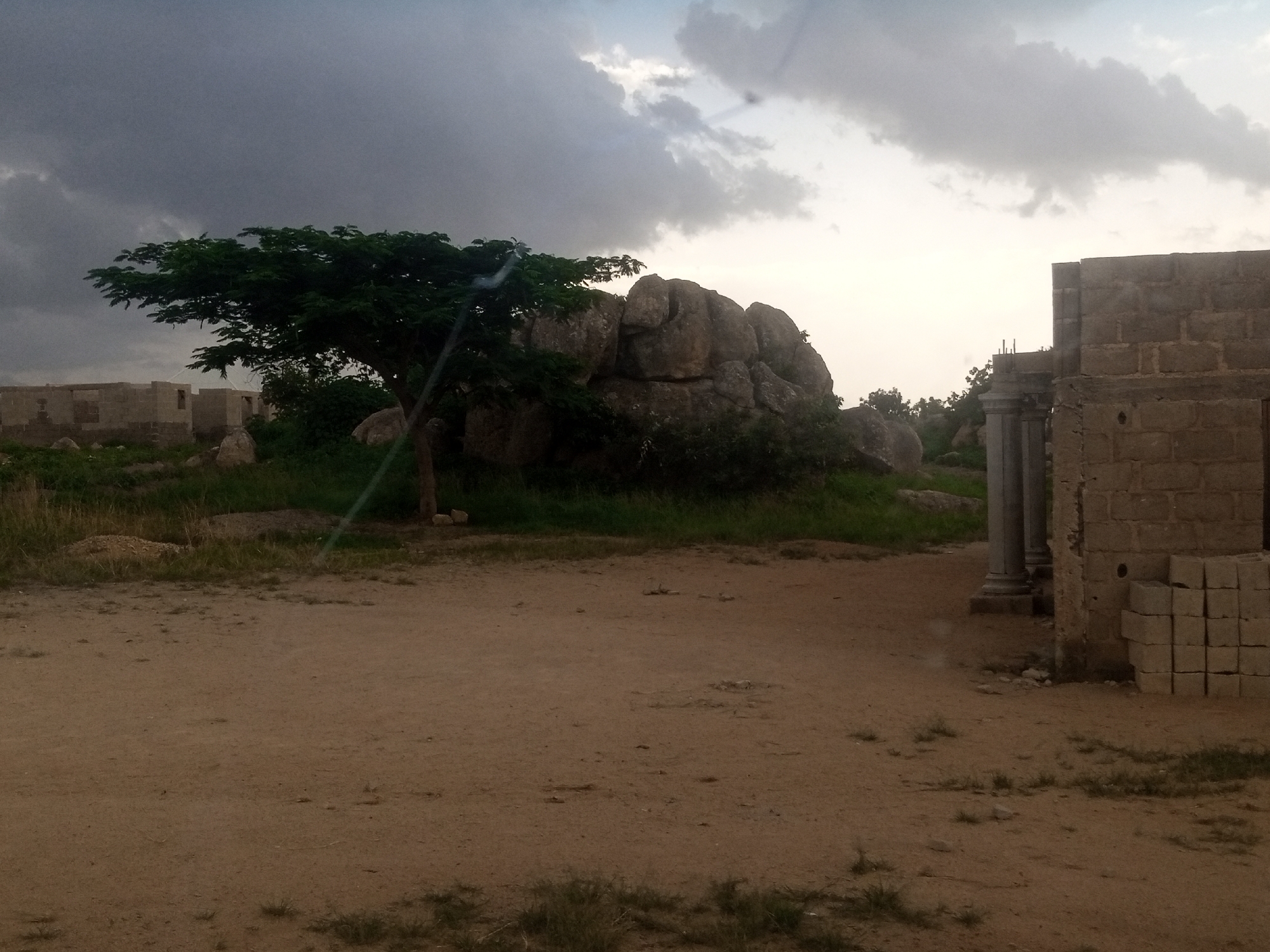 The image depicts an outdoor scene in an undeveloped area, possibly on the outskirts of a city. In the foreground, a dirt path dominates the lower half of the frame, with sparse dry grass and scattered small stones. To the left, the construction of several buildings is underway, with concrete block walls partially erected and exposed rebar. A single tree with green foliage stands prominently in the mid-ground, its trunk supported by a wooden stake. Behind the tree, a cluster of large, grey boulders forms a natural formation. To the right of the frame, another partially constructed building is visible, made of concrete blocks, with a stack of cement blocks in front of it. Several cylindrical pillars, appearing to be part of a larger structure, are also present. The sky is overcast with dramatic grey clouds, suggesting impending or recent rain. The overall impression is one of a rural or semi-urban environment in a state of development or disrepair, with elements of natural landscape and human construction.