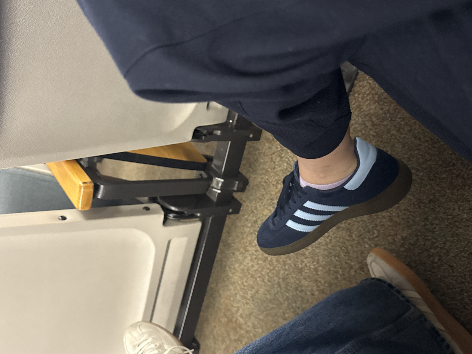 The image shows a person seated on what appears to be public transportation, with another individual's legs and feet visible nearby. The seat is a light gray color, with metal framing. The person in focus wears dark blue athletic shoes with light blue stripes and dark blue pants. The second person wears denim jeans and white/beige sneakers. The setting is an interior with a carpeted floor, likely within a bus or train in an unknown location. The location in San Antonio, United States of America, cannot be determined.