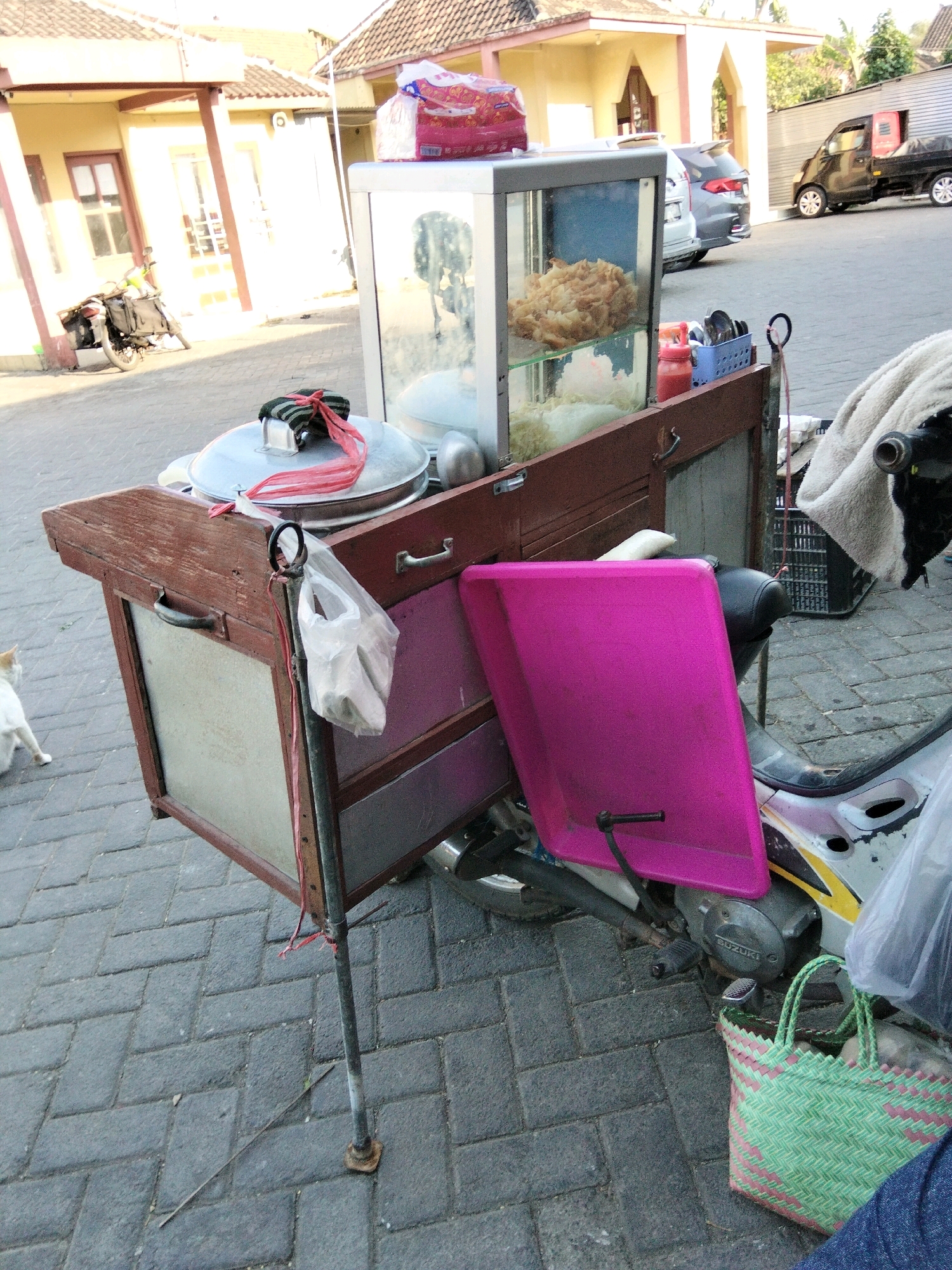 The image depicts a street scene in Banyudono, Indonesia, likely a commercial or residential area. The primary subject is a food cart, modified from a motorcycle, positioned on a paved walkway. The cart is constructed of wood and metal, featuring a glass display case filled with food items. A large pink plastic container is attached to the side of the cart, and a green woven bag sits on the ground nearby. To the left of the cart, a white cat is visible, walking away from the camera. In the background, a building with yellow walls and tiled roofs stands, and a dark-colored truck is parked further down the street. The overall environment suggests an outdoor setting, possibly in the early morning or late afternoon due to the lighting.