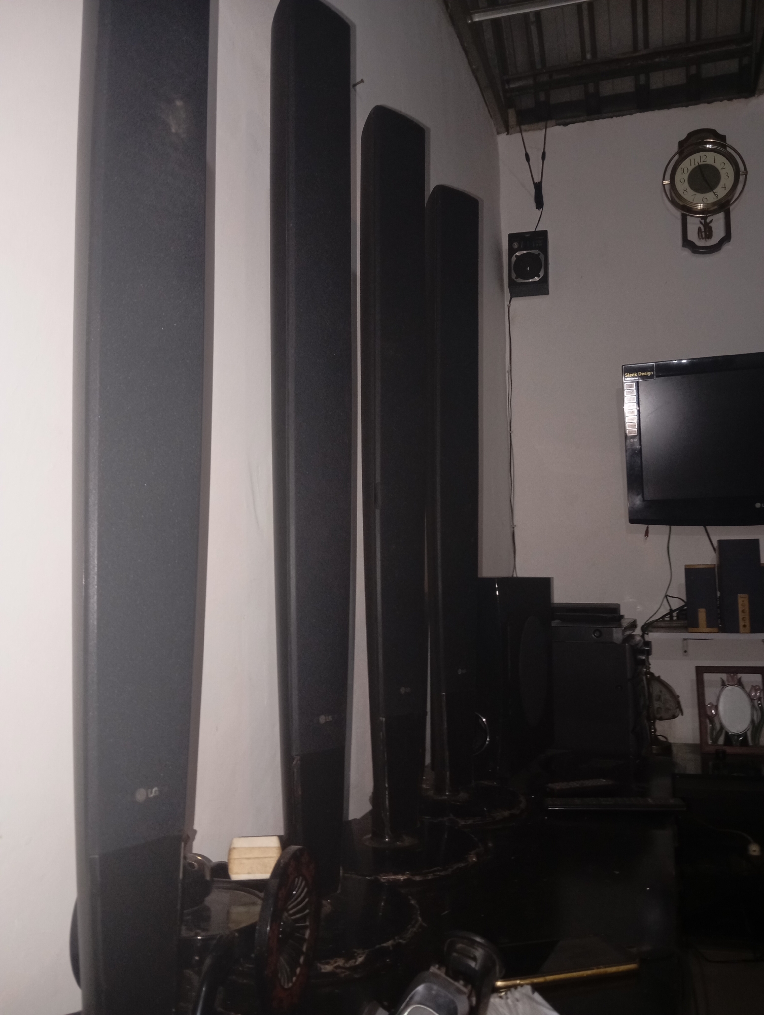 In a room in Kedungbenda, Indonesia, a collection of tall, dark speakers stands against a white wall. The speakers are arranged in a row, with the leftmost speaker being the widest. Some of the speakers have the LG logo visible. In the background, a television is mounted on the wall, and a round clock with a pendulum hangs above it. Various electronic devices and accessories are placed on shelves and surfaces around the room. The lighting appears to be dim, casting shadows and creating a somewhat cluttered atmosphere. The ceiling is visible in the upper portion of the image, appearing to be made of corrugated metal.