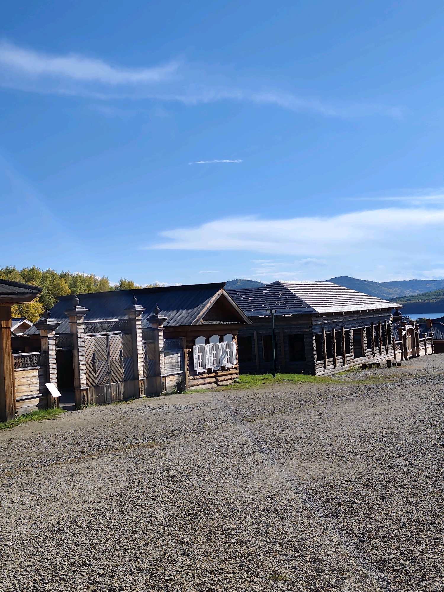 The image depicts a rustic scene at the architectural-ethnographic museum "Taltsy" in the Irkutsk Oblast of Russia. The foreground is dominated by a gravel path leading towards a cluster of wooden buildings that appear to be historical structures. These buildings feature traditional Russian architecture, with log construction, wooden gates, and decorative window frames. The main building in the center is a long, low structure with an unfinished roof, suggesting ongoing restoration or preservation work.
Behind the buildings, a line of trees with autumn foliage is visible, indicating the season. Further in the distance, rolling hills and a body of water, likely Lake Baikal, can be seen under a bright blue sky with scattered white clouds. The overall impression is one of a preserved historical site set within a natural, scenic environment. There are no people visible in the image.